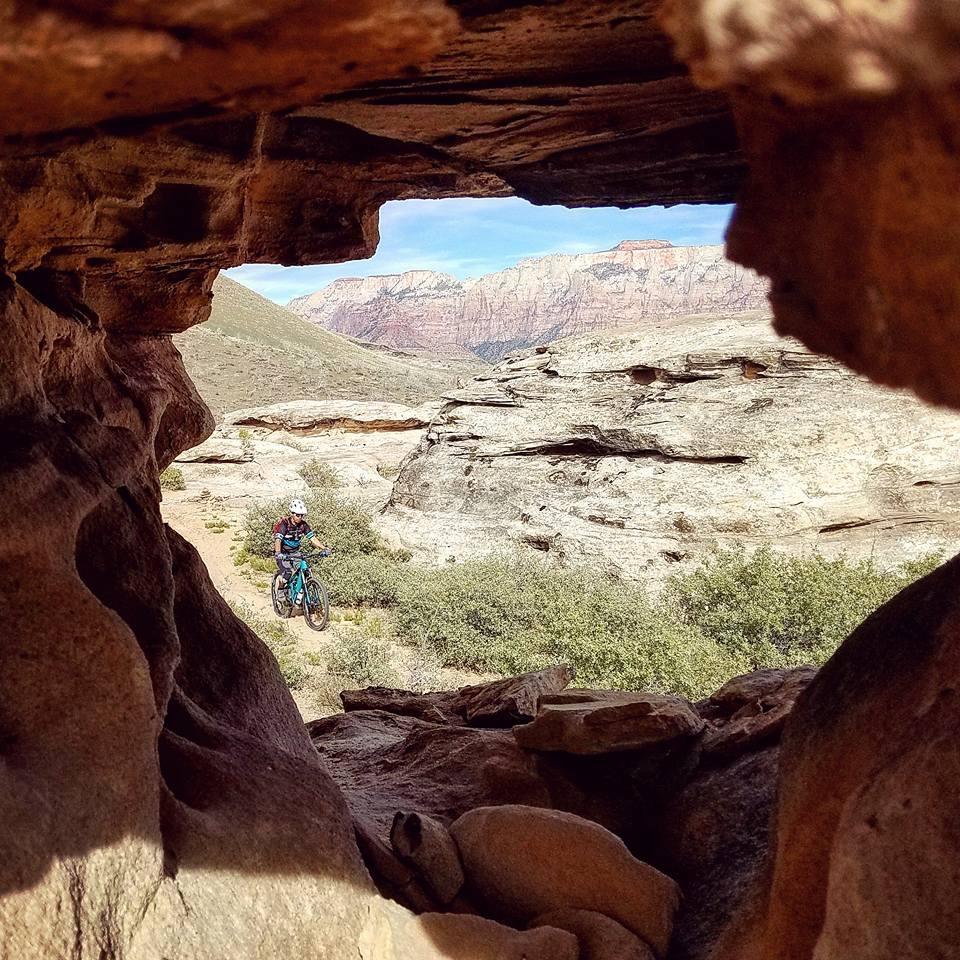 A mountain biker rides through a rocky landscape, viewed through a natural rock formation that frames the scene. In the background, hills and colorful cliffs rise against a blue sky, showcasing the beauty of the outdoor terrain. Guacamole Mesa mountain bike trail.
