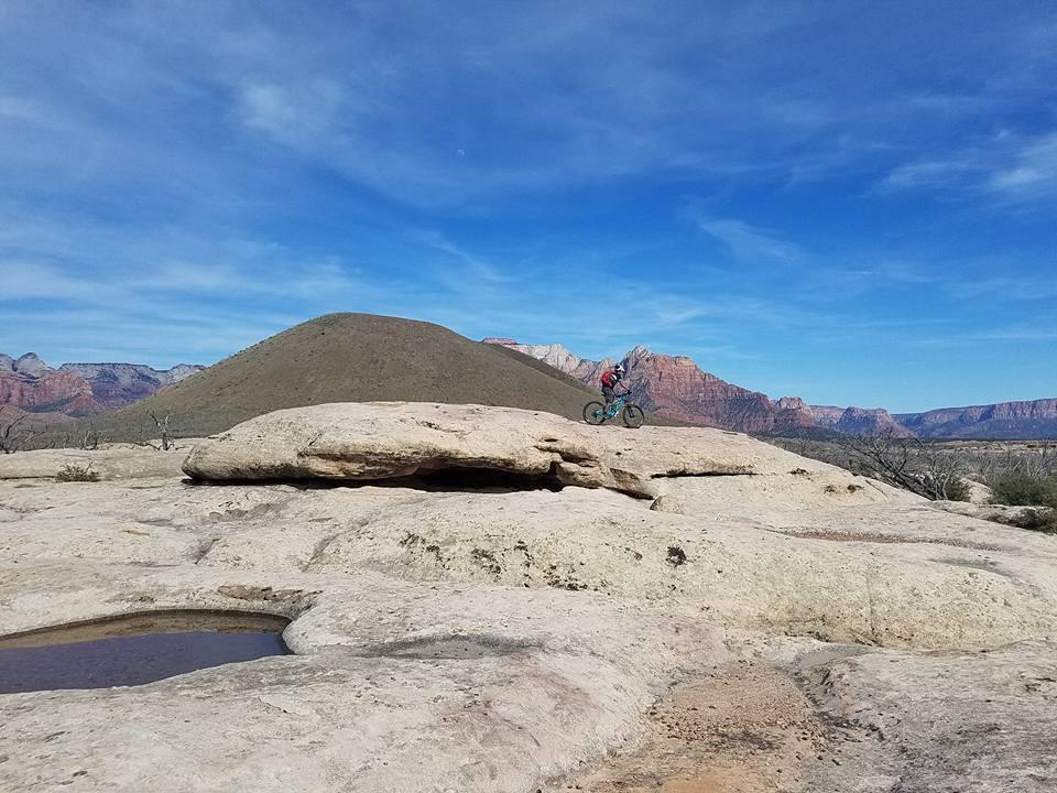A mountain biker navigating rocky terrain on a large flat boulder, with a backdrop of colorful mountains and a clear blue sky. Sparse vegetation is visible in the foreground, and a small pool of water can be seen on the rock surface. Guacamole Mesa mountain bike trail.
