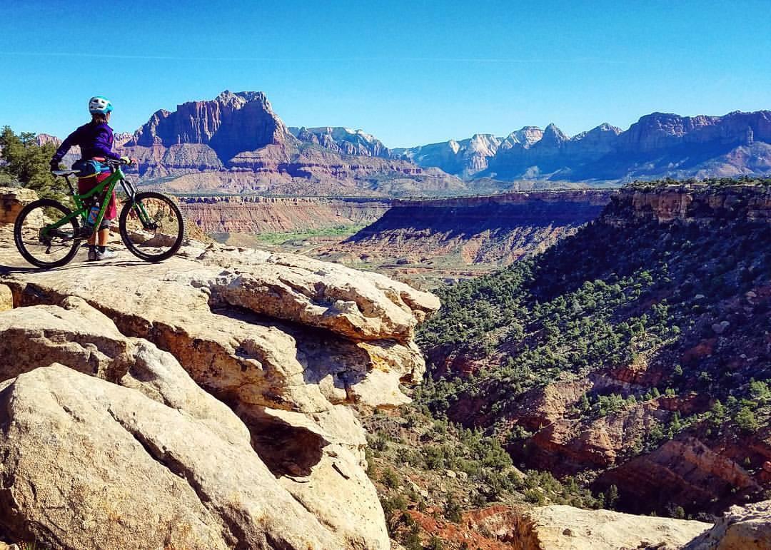 A mountain biker stands on a rocky outcrop overlooking a vast valley, surrounded by stunning mountain scenery under a clear blue sky. The biker is dressed in colorful attire with a helmet and is positioned next to a green mountain bike. The landscape features layered cliffs, greenery, and distant mountains with snow-capped peaks. Wire Mesa Loop mountain bike trail.