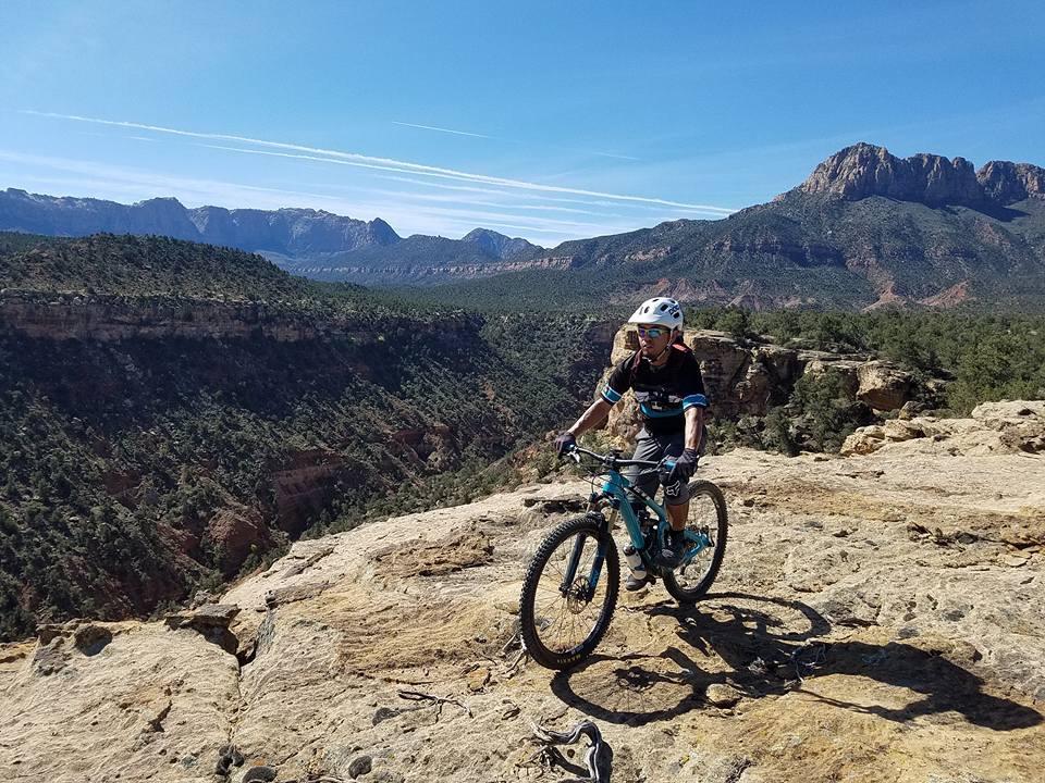 A mountain biker wearing a helmet and sunglasses rides on a rocky terrain with a scenic view of mountains and valleys in the background. The sky is clear and blue, highlighting the natural landscape. Wire Mesa Loop mountain bike trail.