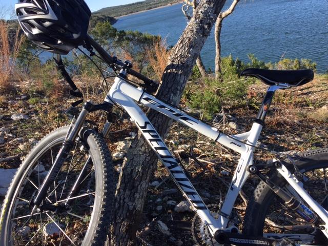 Specialized Epic: A close-up view of a white mountain bike resting against a tree with a body of water in the background. The bike is equipped with a helmet mounted on the handlebars, and the surrounding area features rocks and greenery, suggesting a scenic outdoor setting.