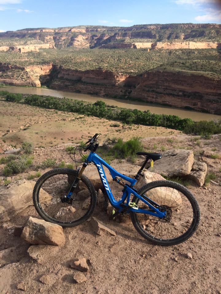 Pivot Mach 429 Carbon: Mountain bike resting on rocky terrain, with a scenic view of a river meandering through a canyon and rugged cliffs in the background. The landscape features earthy tones and green vegetation, indicating a natural outdoor setting.