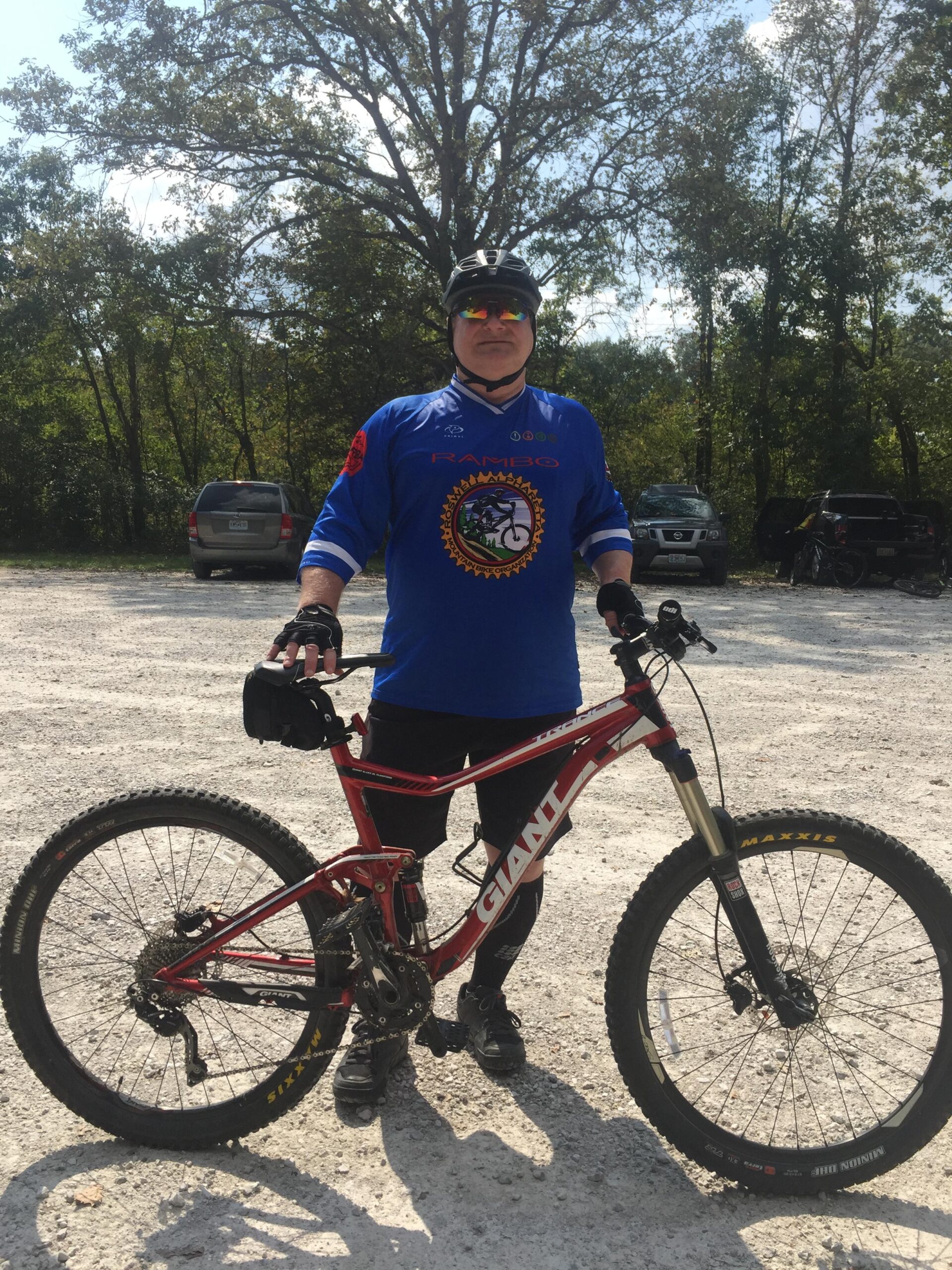 Giant Trance: A person standing next to a red mountain bike, wearing a blue cycling jersey with the word "RAMBO" on it, black shorts, and a helmet. The background features trees and parked vehicles on a gravel surface.