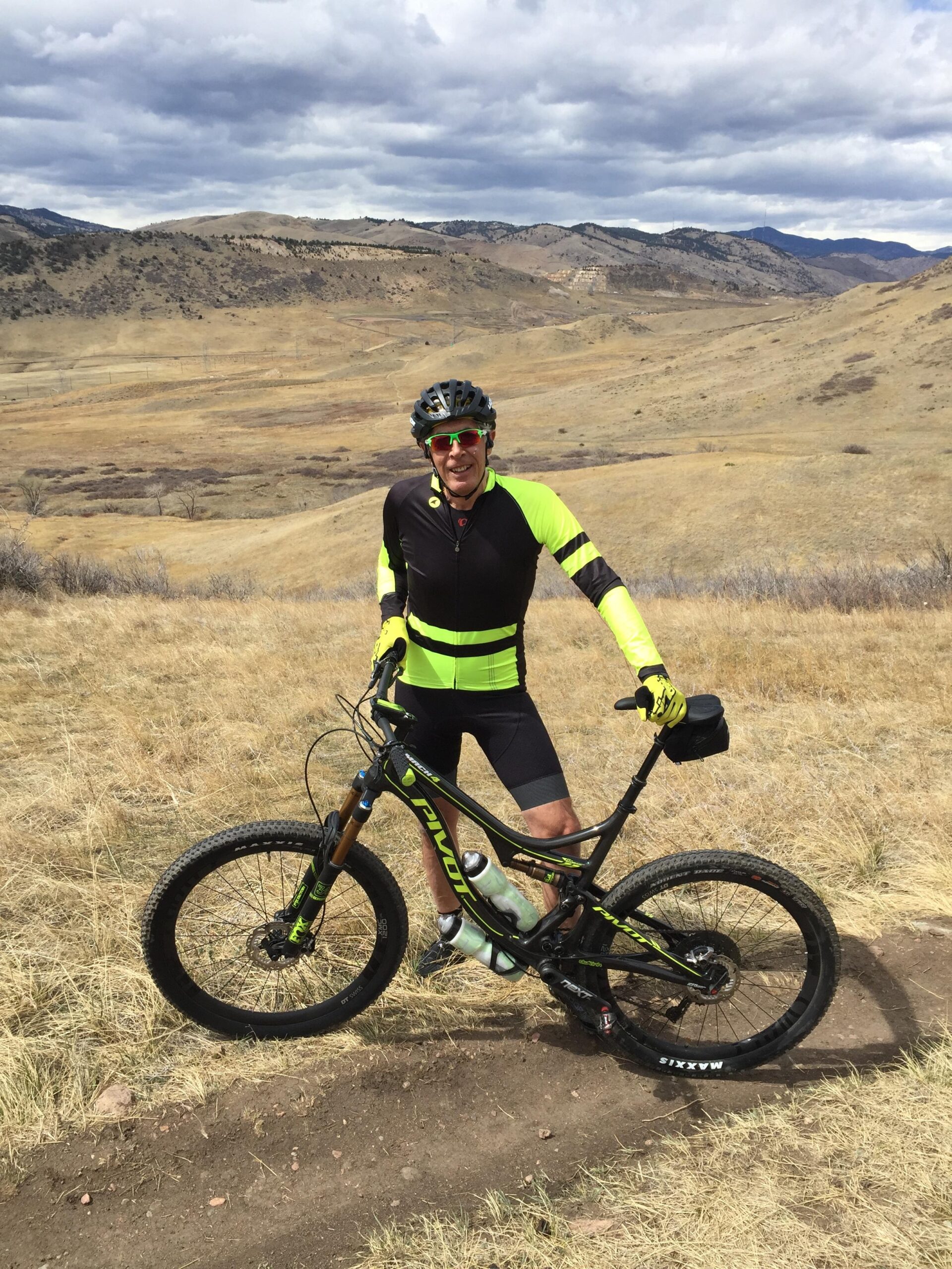 Pivot Mach 4: A cyclist dressed in a bright yellow and black outfit stands next to a mountain bike on a trail, surrounded by a hilly, grassy landscape under a cloudy sky.