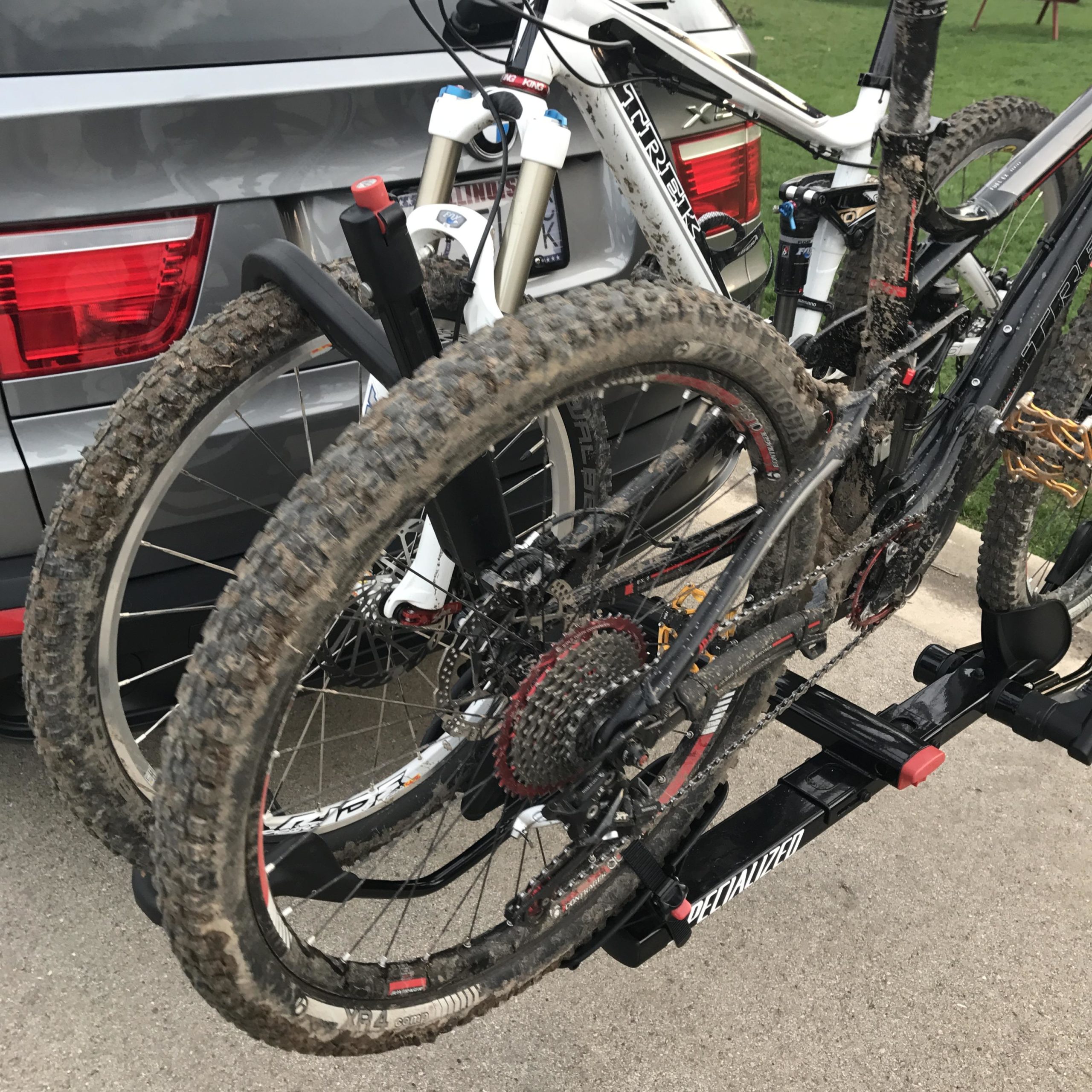 Two mountain bikes mounted on a bike rack attached to the back of a vehicle. The bikes are covered in mud, indicating recent outdoor activity. One bike has a black frame with red and gold accents, while the other features a white and black frame. The background includes part of the car’s rear and green grass. Petrifying Springs mountain bike trail.