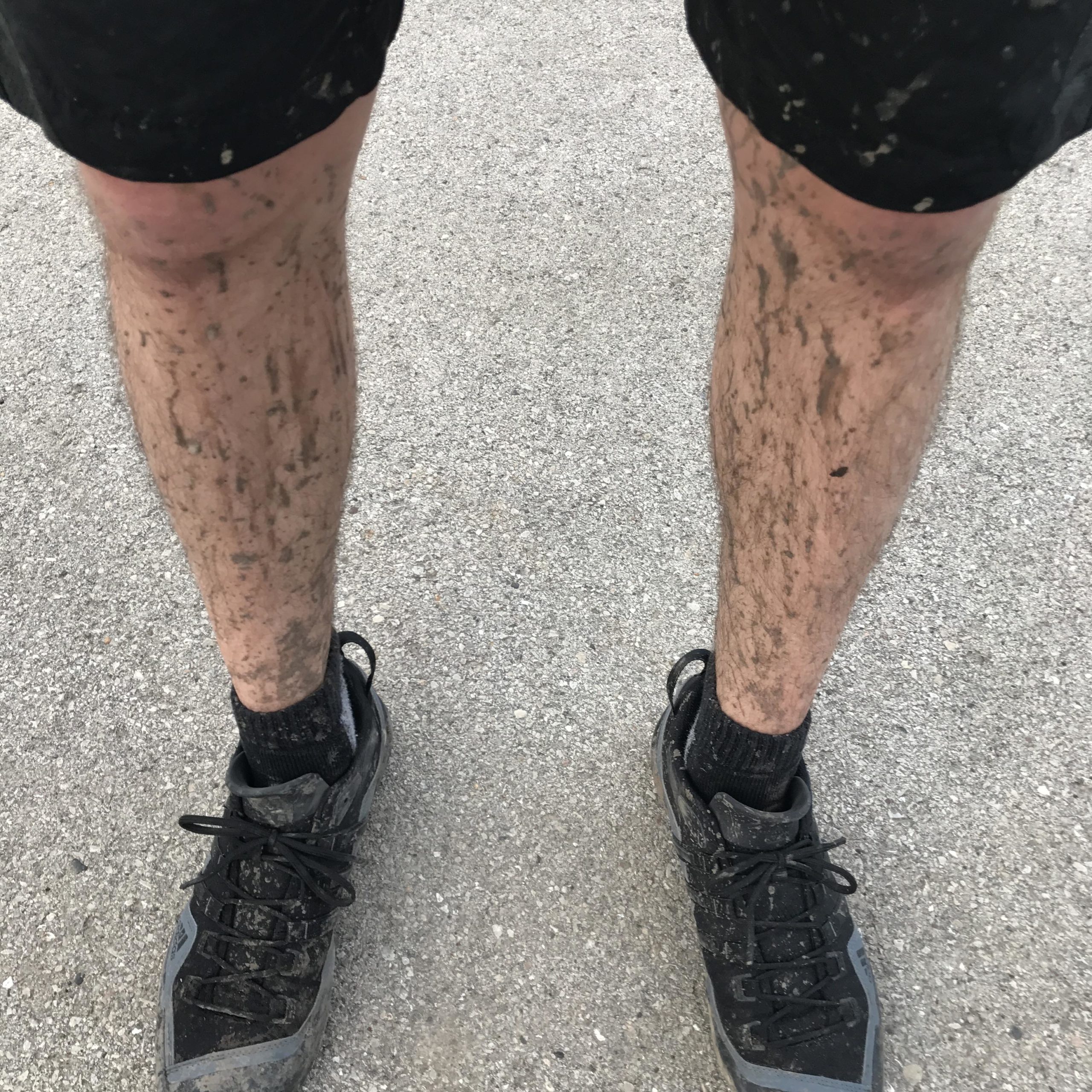 A pair of muddy legs wearing black shorts, with visible dirt and grime from outdoor activities, stand on a concrete surface. The person is wearing black athletic shoes and socks. Petrifying Springs mountain bike trail.