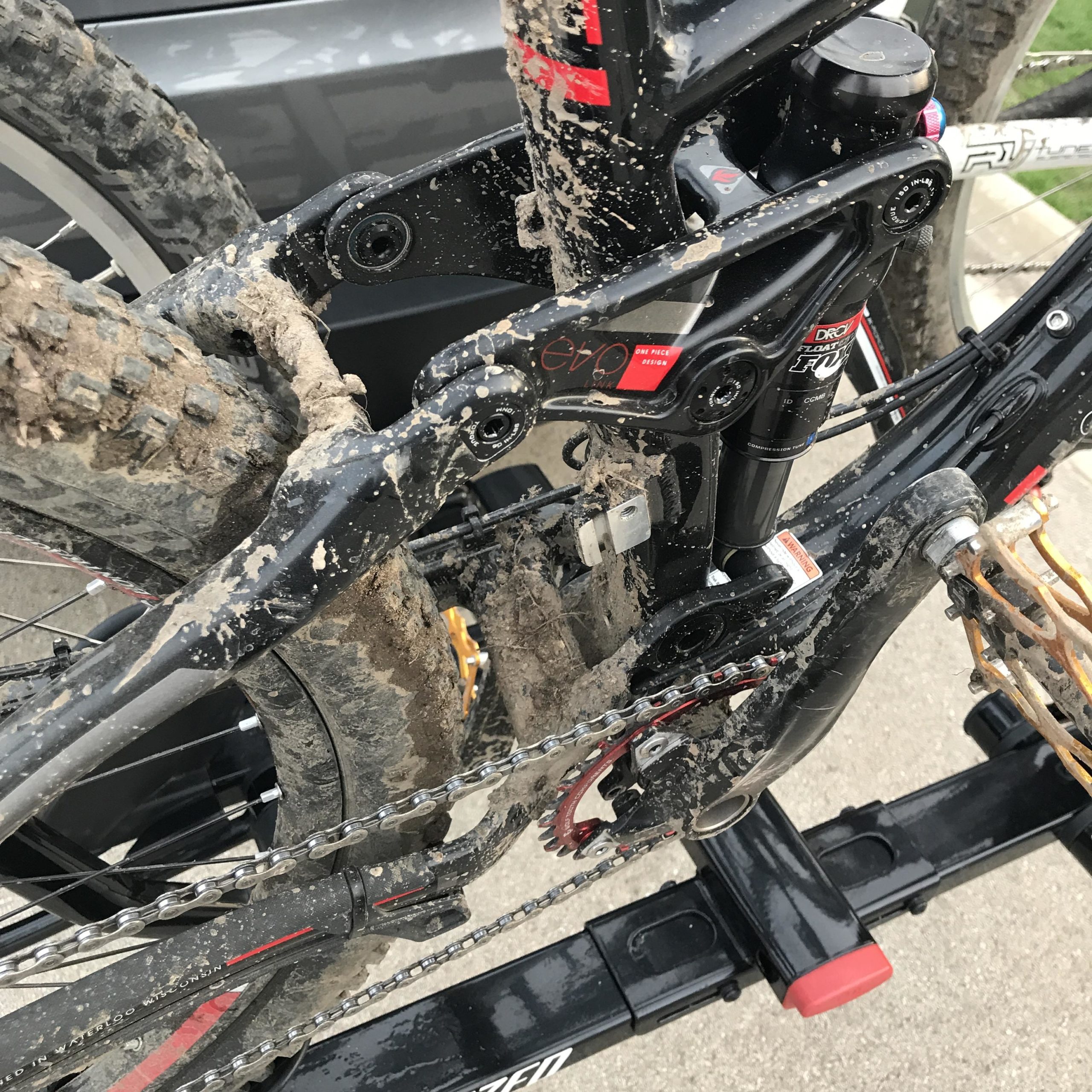 Close-up view of a muddy mountain bike's rear suspension and drivetrain components, highlighting dirt and grime accumulated on the frame and parts after a ride. The image shows the chain, gears, and shock absorber, with a focus on the wear from outdoor cycling conditions. Petrifying Springs mountain bike trail.