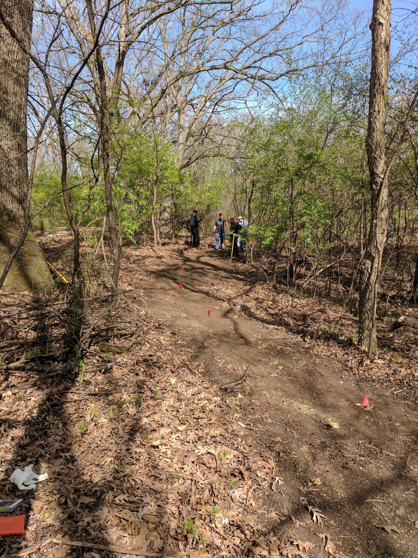 A trail in a wooded area with bare trees and budding greenery, where three people are gathered near a dirt path marked with small red flags. Leaf litter covers the ground, and there are signs of recent activity. The sky is clear and blue, indicating a sunny day. Raceway Woods mountain bike trail.