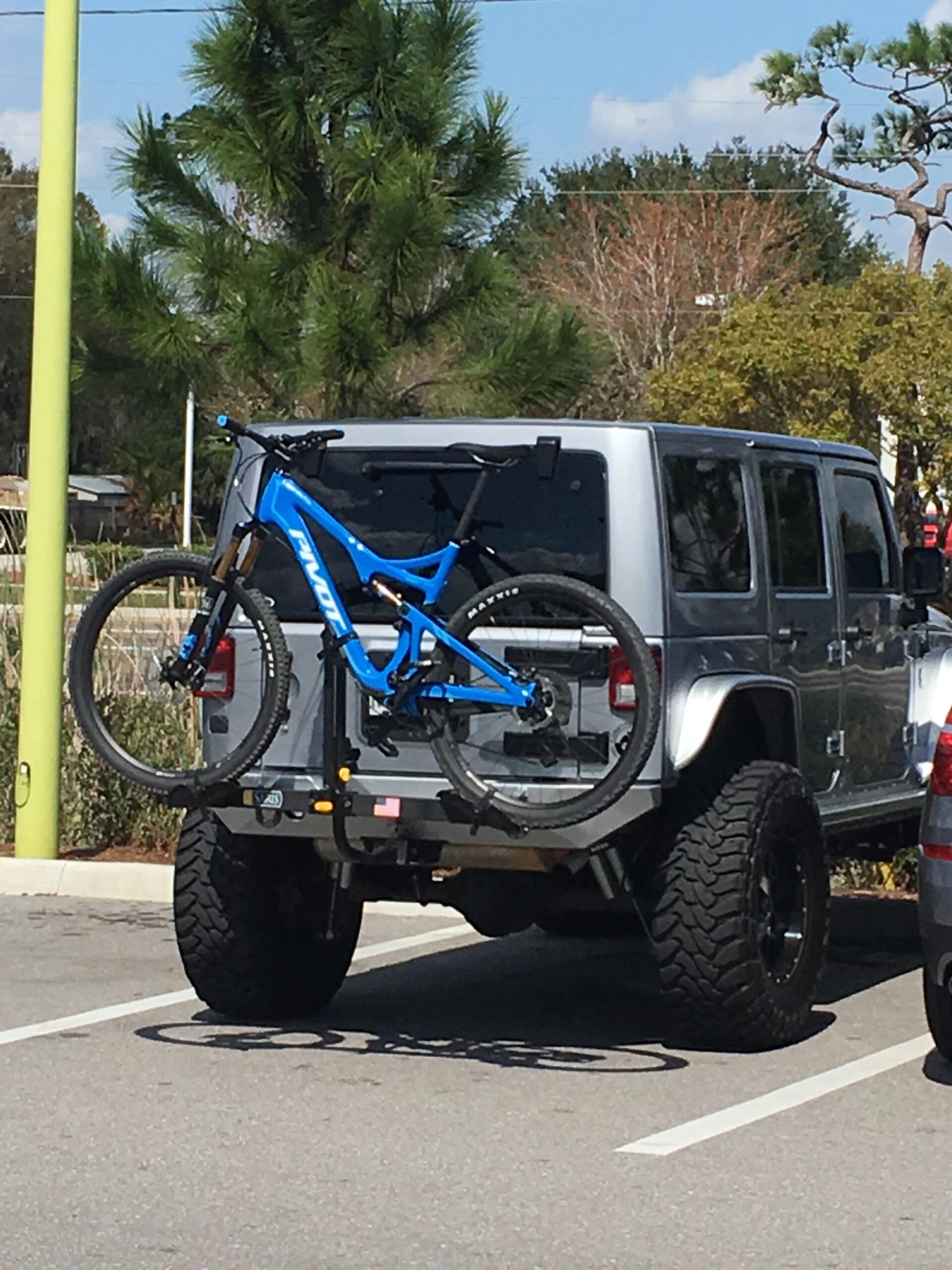 Pivot Mach 429 Carbon: A silver SUV parked in a parking lot with a blue mountain bike mounted on a bike rack at the rear. Surrounding the vehicle are various trees and a clear blue sky.