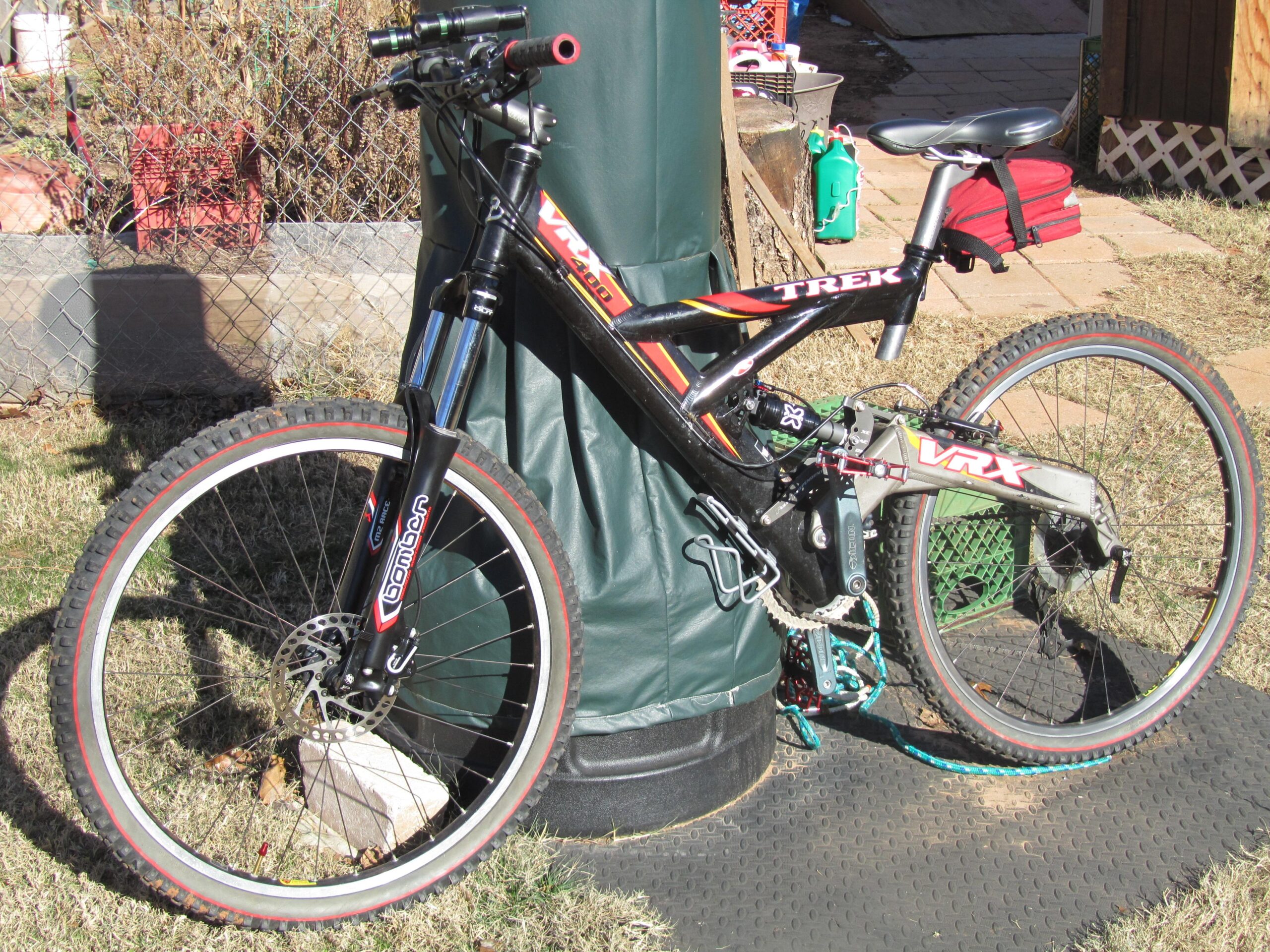 Giant XTC 29er 2: A black and red Trek mountain bike leaning against a green cover, featuring a suspension fork and decorated wheels with red accents. The bike is positioned on a textured black mat, surrounded by a grassy area and various outdoor elements in the background.