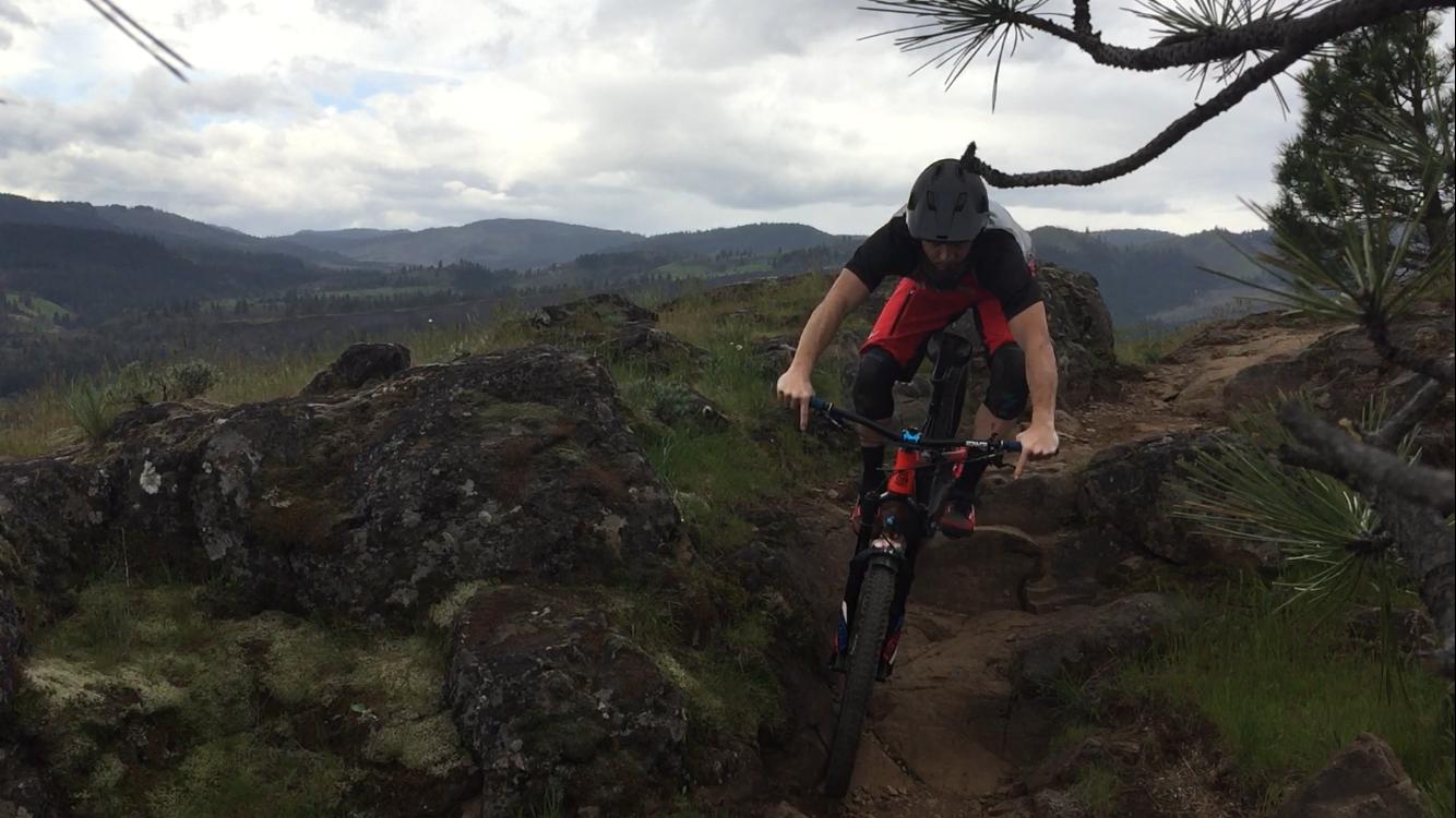 A mountain biker in protective gear navigates a rocky trail surrounded by greenery and distant hills under a cloudy sky. Syncline mountain bike trail.