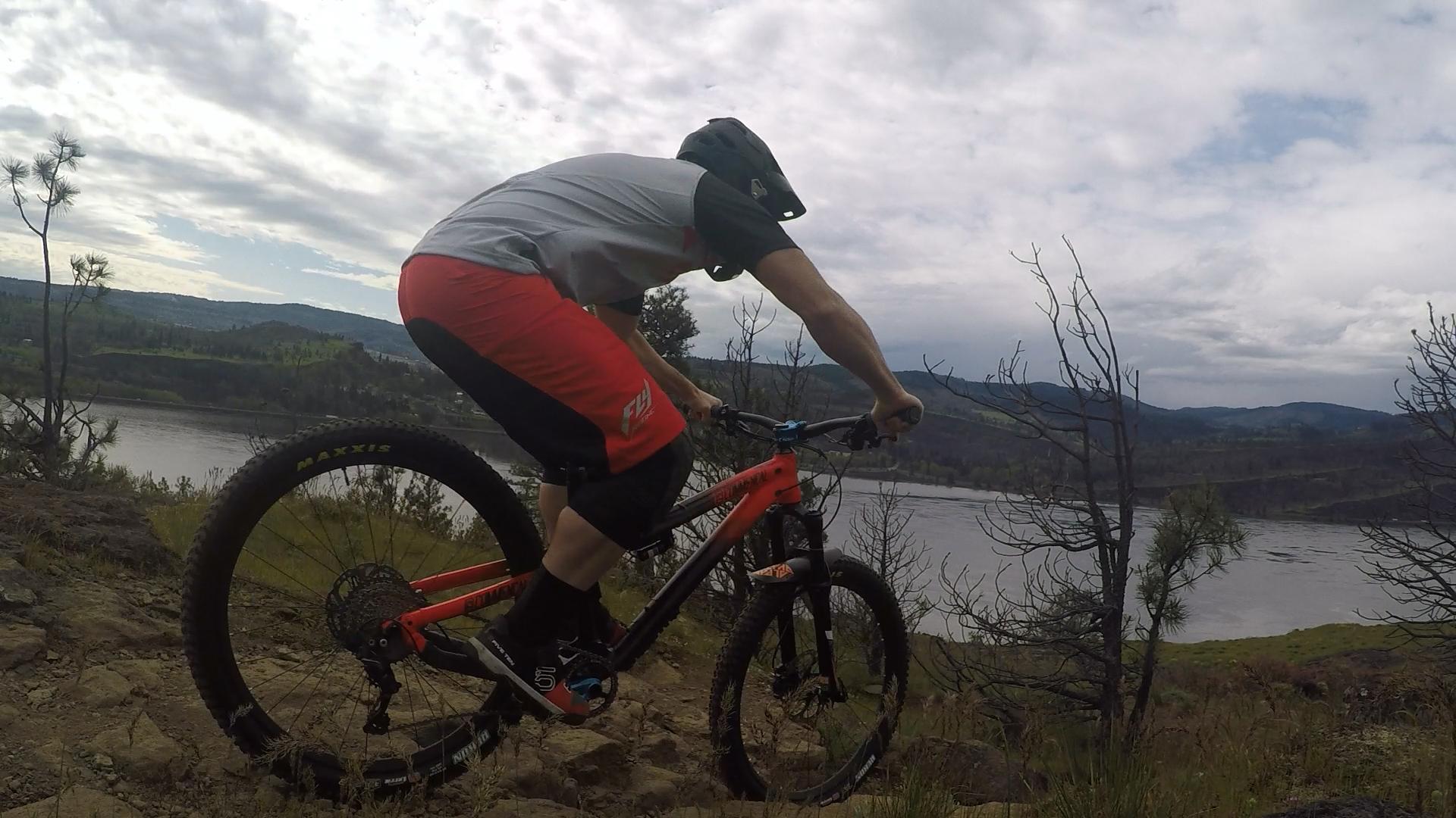 A cyclist in a gray and red outfit rides a mountain bike on rocky terrain near a river, with hills and trees in the background under a cloudy sky. Syncline mountain bike trail.
