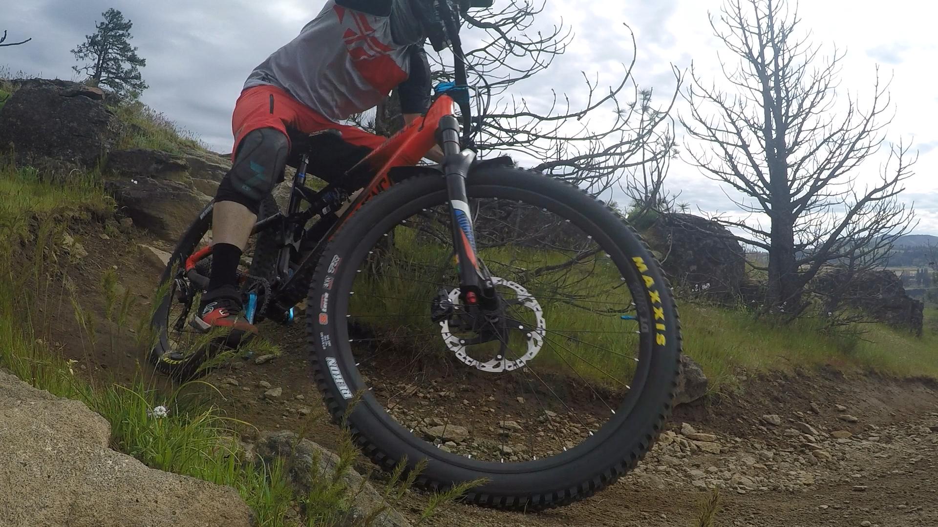 A cyclist in a red and gray jersey is navigating a rocky mountain biking trail, with one wheel lifted off the ground. The surrounding landscape features grassy areas, scattered rocks, and sparse trees under a cloudy sky. Syncline mountain bike trail.