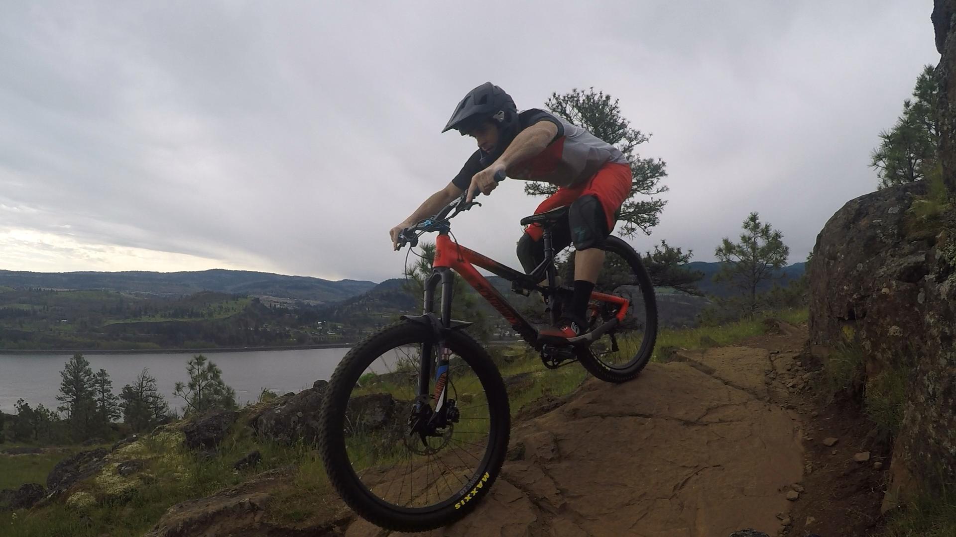 A mountain biker performing a jump on rocky terrain, showcasing a dynamic pose while riding a red bike. The background features a scenic view of hills and a river under a cloudy sky. Syncline mountain bike trail.