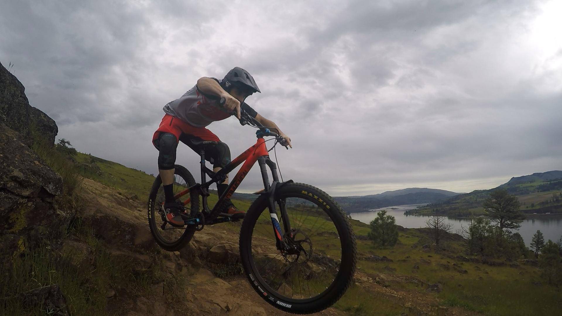 A mountain biker in a helmet and protective gear rides over rocky terrain with a scenic view of a river and hills in the background. The sky is overcast, adding to the adventurous atmosphere. Syncline mountain bike trail.