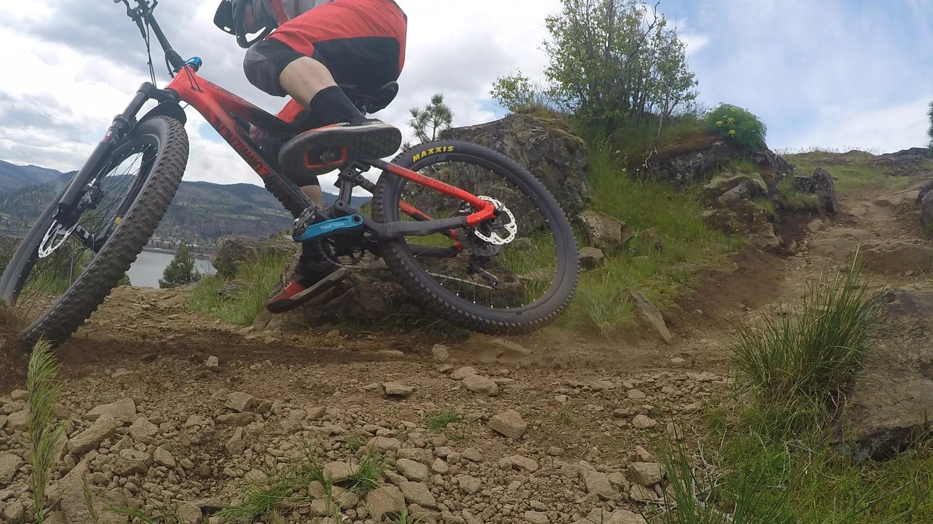 A mountain biker navigating a rocky trail, kicking up dirt as they make a sharp turn. The bike features large tires and a vibrant red and black frame, with a scenic landscape of hills and a lake in the background under a partly cloudy sky. Syncline mountain bike trail.