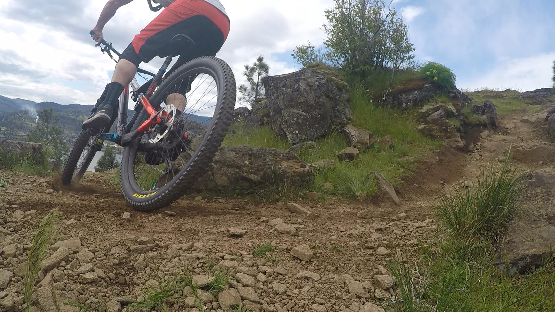 A close-up view of a mountain biker navigating a rocky dirt trail, showcasing the bike's back wheel kicking up dust as it takes a turn. The background features a lush green landscape with trees and distant hills under a partly cloudy sky. Syncline mountain bike trail.