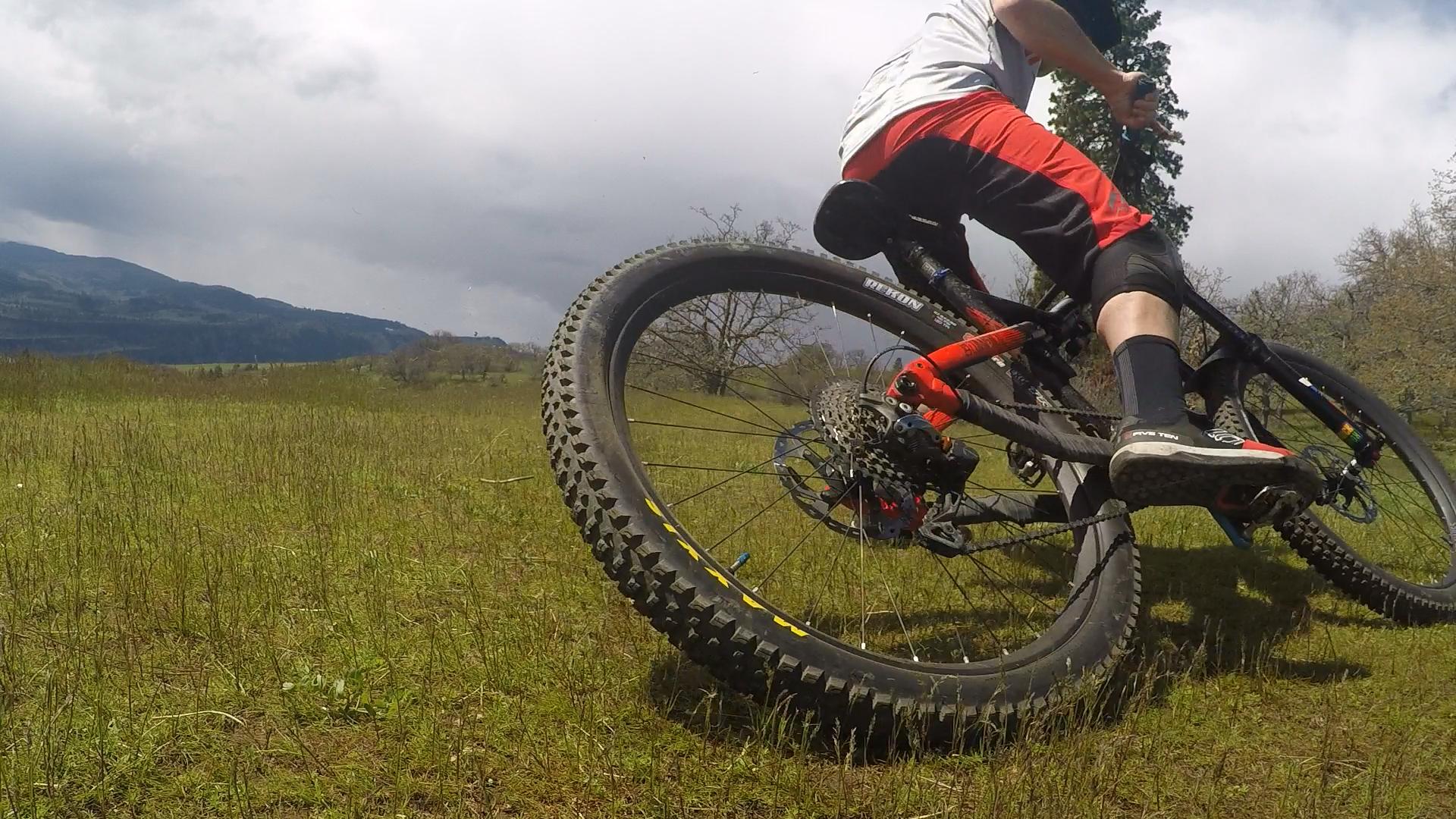 A mountain biker maneuvering on a grassy field, with the bike's rear wheel prominently featured. The cyclist is wearing red shorts and a grey shirt, showcasing dynamic movement against a backdrop of cloudy skies and distant mountains. Syncline mountain bike trail.
