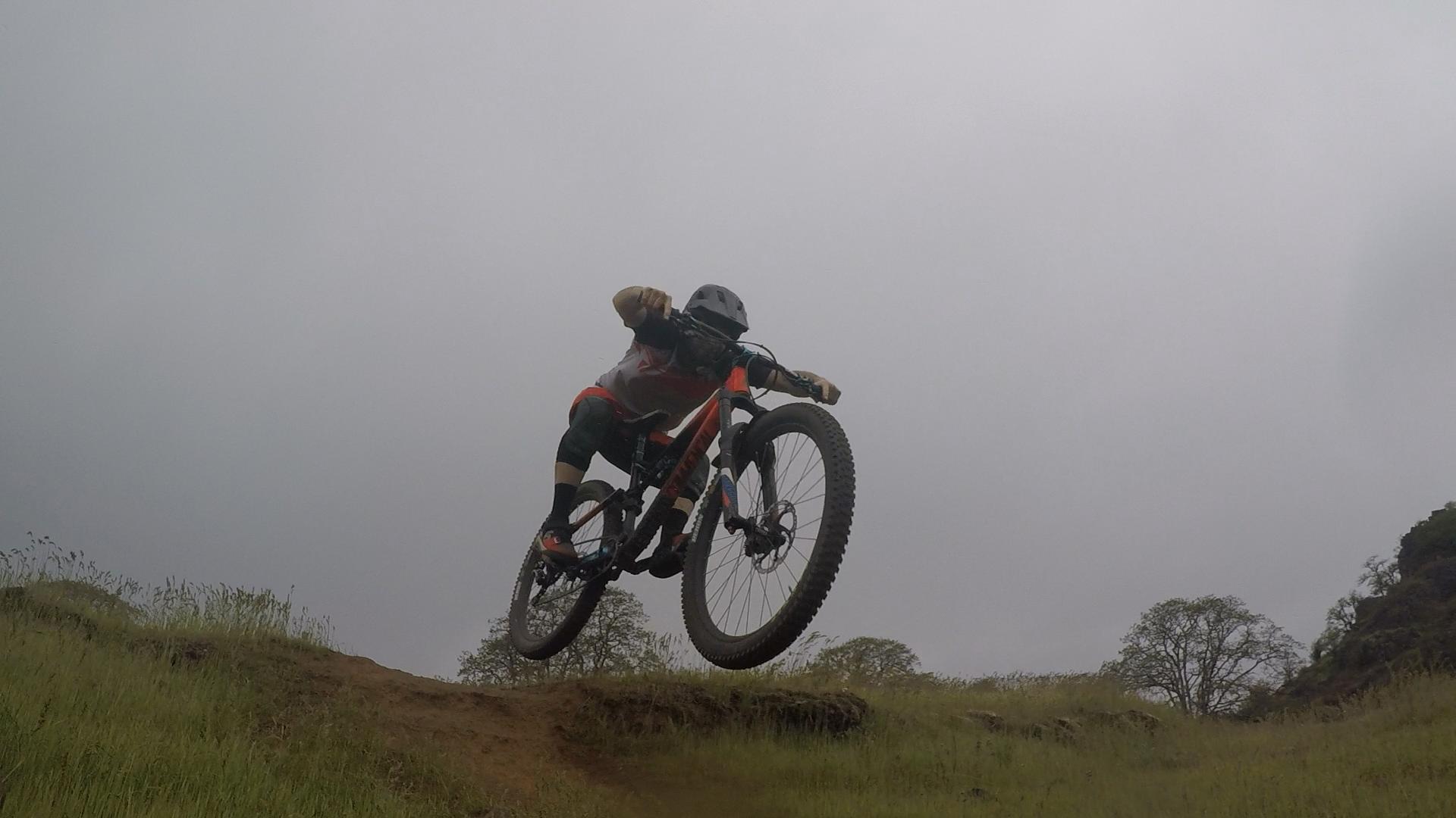 A mountain biker performing a jump over a dirt ramp on a cloudy day, surrounded by tall grass and distant trees. Syncline mountain bike trail.