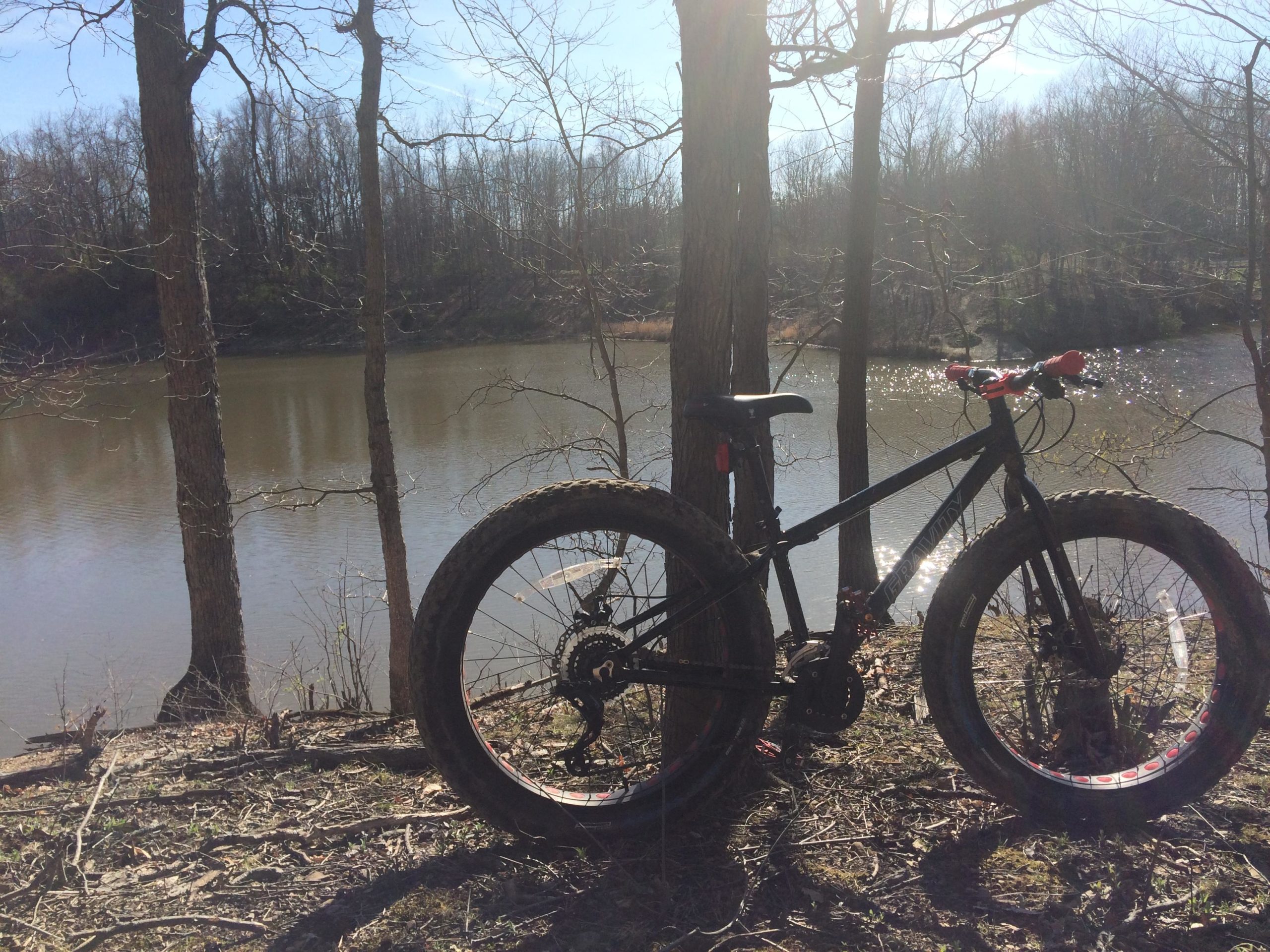A black fat tire bicycle leaning against a tree near a calm lake, surrounded by bare trees and sunlight reflecting off the water. Alum Creek Phase II mountain bike trail.