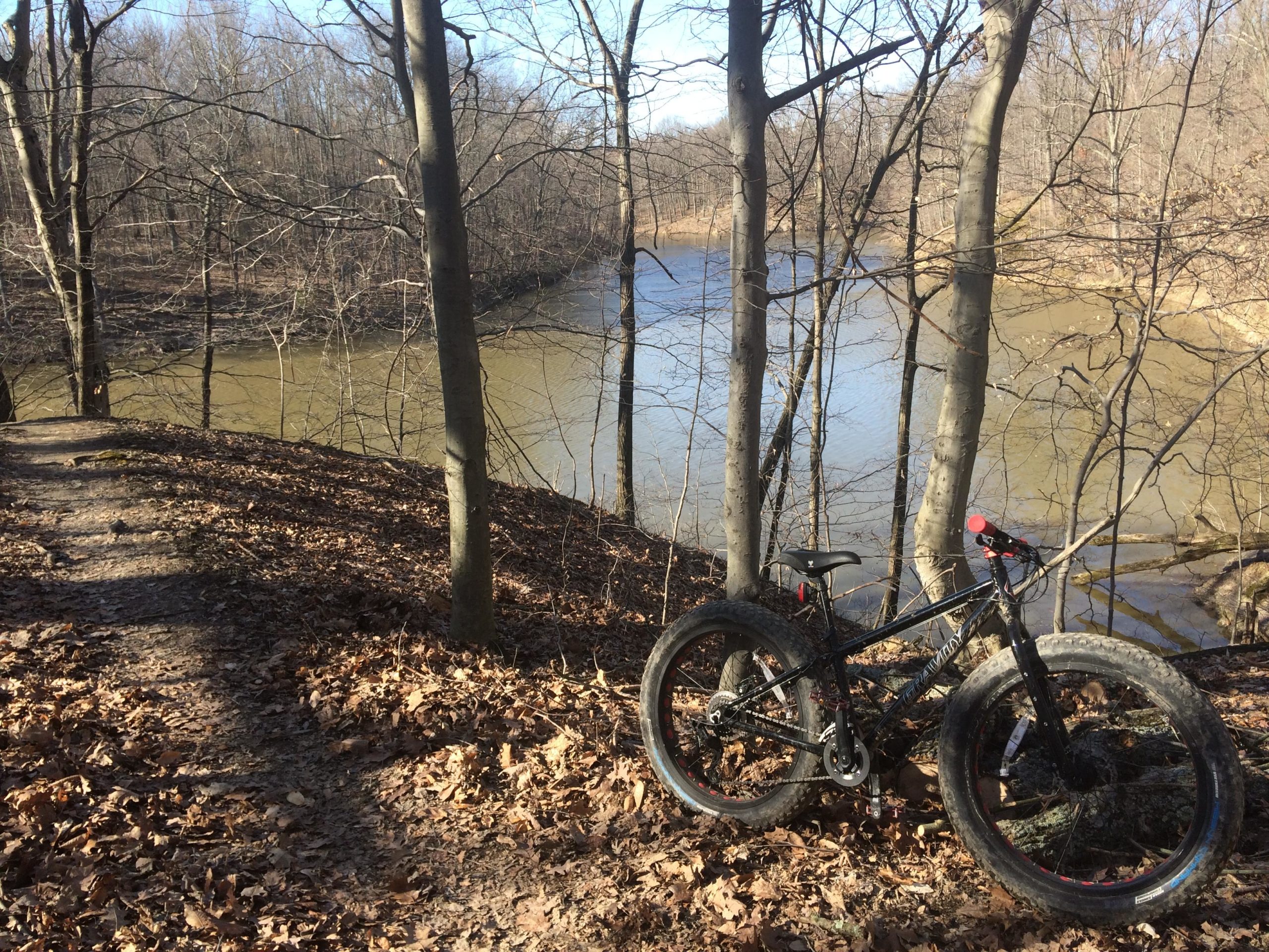 A fat tire bike resting on a leaf-covered trail next to a calm river, surrounded by leafless trees and a clear blue sky. Alum Creek Phase II mountain bike trail.