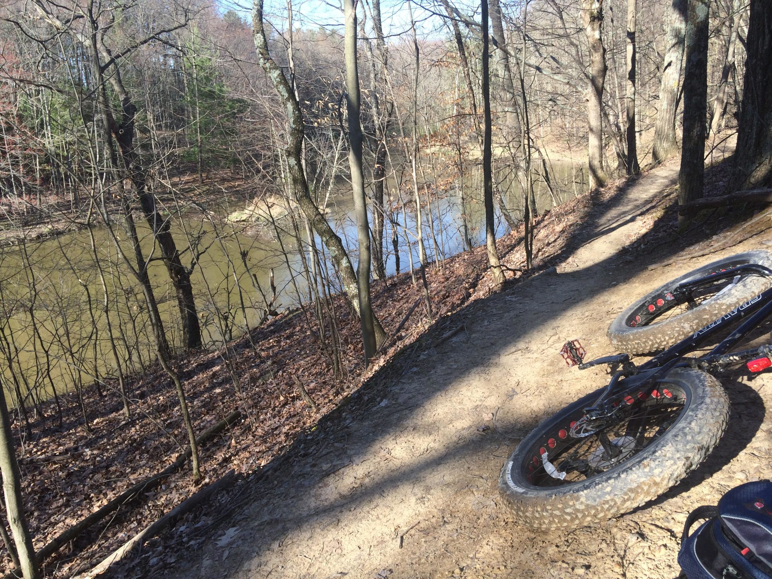 A mountain bike laying on a dirt trail beside a calm river, surrounded by bare trees and shrubs in a forested area. The scene is captured in daylight, highlighting the natural landscape and tranquil setting. Alum Creek Phase II mountain bike trail.