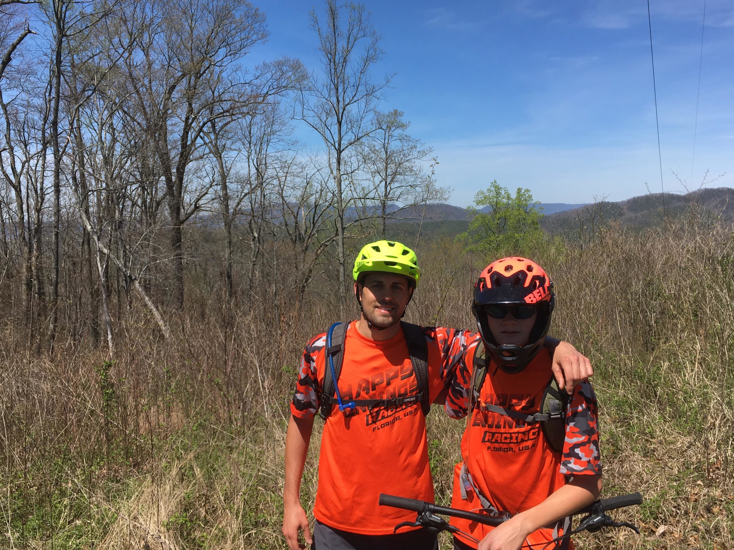 Two mountain bikers posing together in a forested area, wearing bright orange shirts with a racing logo. One biker has a yellow helmet, while the other wears an orange helmet and sunglasses. The background features bare trees and rolling hills under a clear blue sky. Tanasi Trail System mountain bike trail.