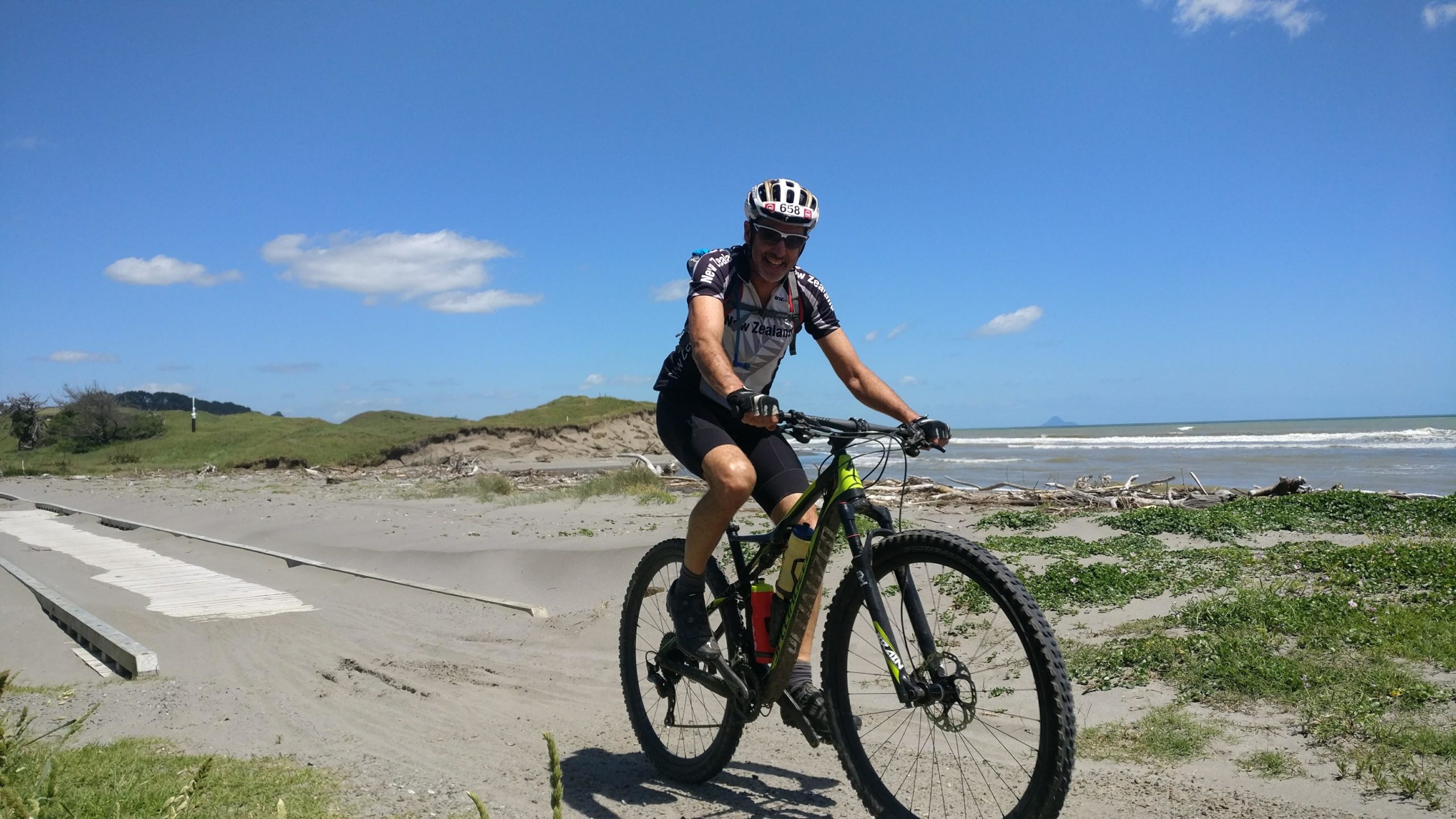 A person riding a mountain bike along a sandy beach with vibrant blue skies and gentle clouds above. The ocean is visible in the background, with waves lapping at the shore. The area has grassy dunes and some driftwood scattered on the beach. Dunes Trail mountain bike trail.