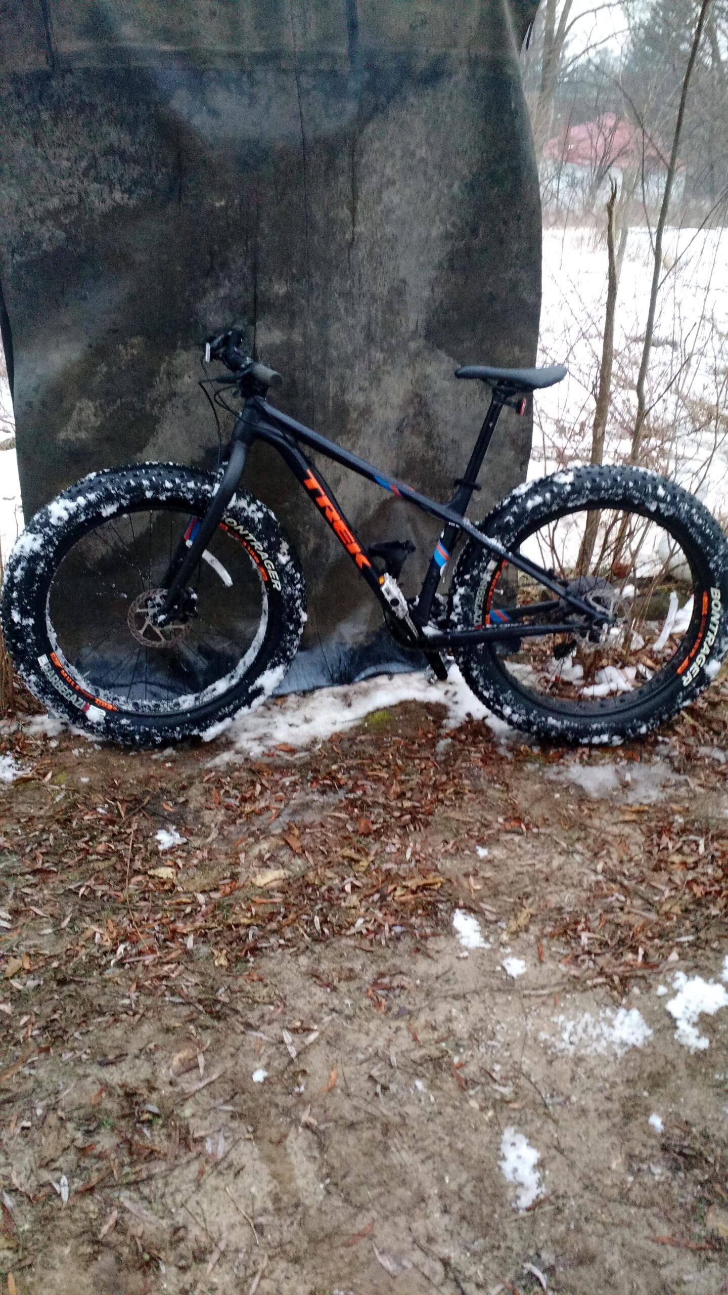 Trek farley 5: A black Trek mountain bike with wide, snow-covered tires parked on a dirt surface surrounded by fallen leaves, near a textured backdrop. Snow patches are visible on the ground, indicating a winter setting.