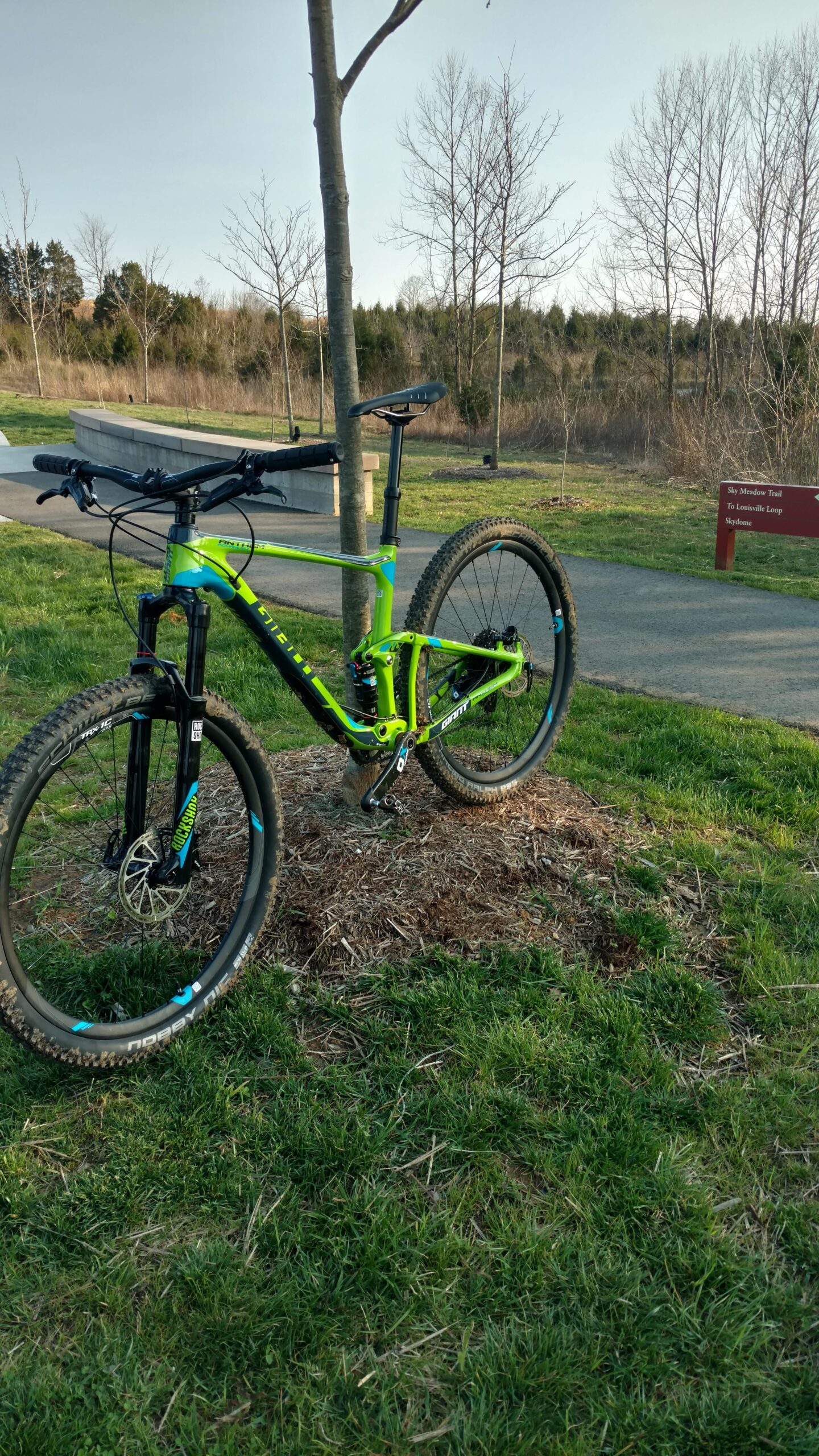 Giant Anthem SX 27.5: A brightly colored mountain bike leaning against a young tree in a grassy area, with a paved path and trees in the background. A sign indicating the direction to the "Sky Meadow Trail" and "Louisville Loop" is visible nearby.