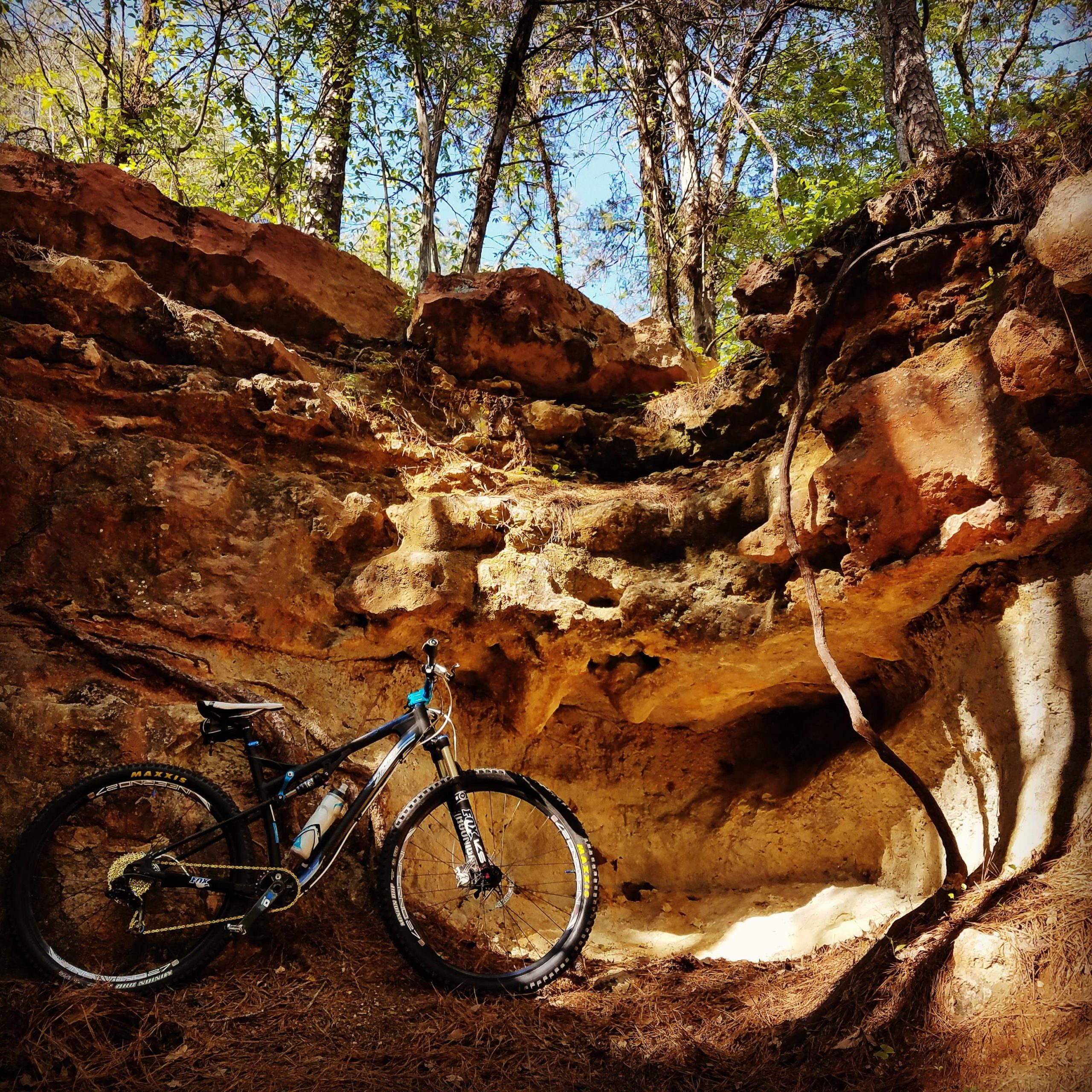 A mountain bike leaning against a rocky outcrop in a forested area, surrounded by trees and dappled sunlight breaking through the foliage. The Ridgeland Trails mountain bike trail.