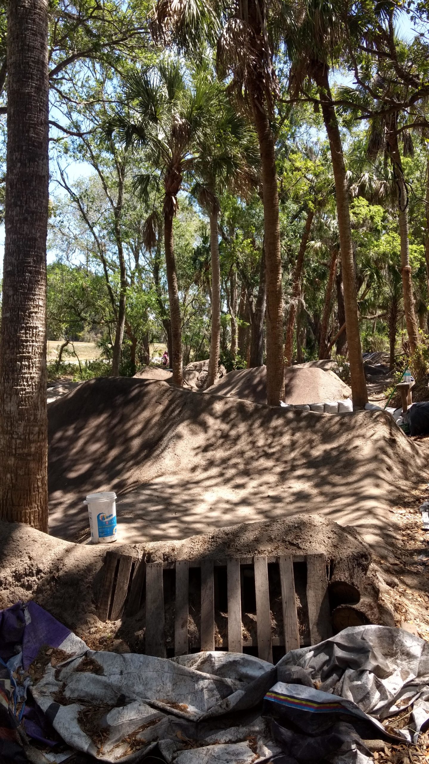 A dirt bike track surrounded by trees, featuring mounds of dirt, a wooden pallet, and a white bucket in the foreground. Sunlight filters through the foliage, creating a dappled light effect on the ground. Grapefruit Trail mountain bike trail.
