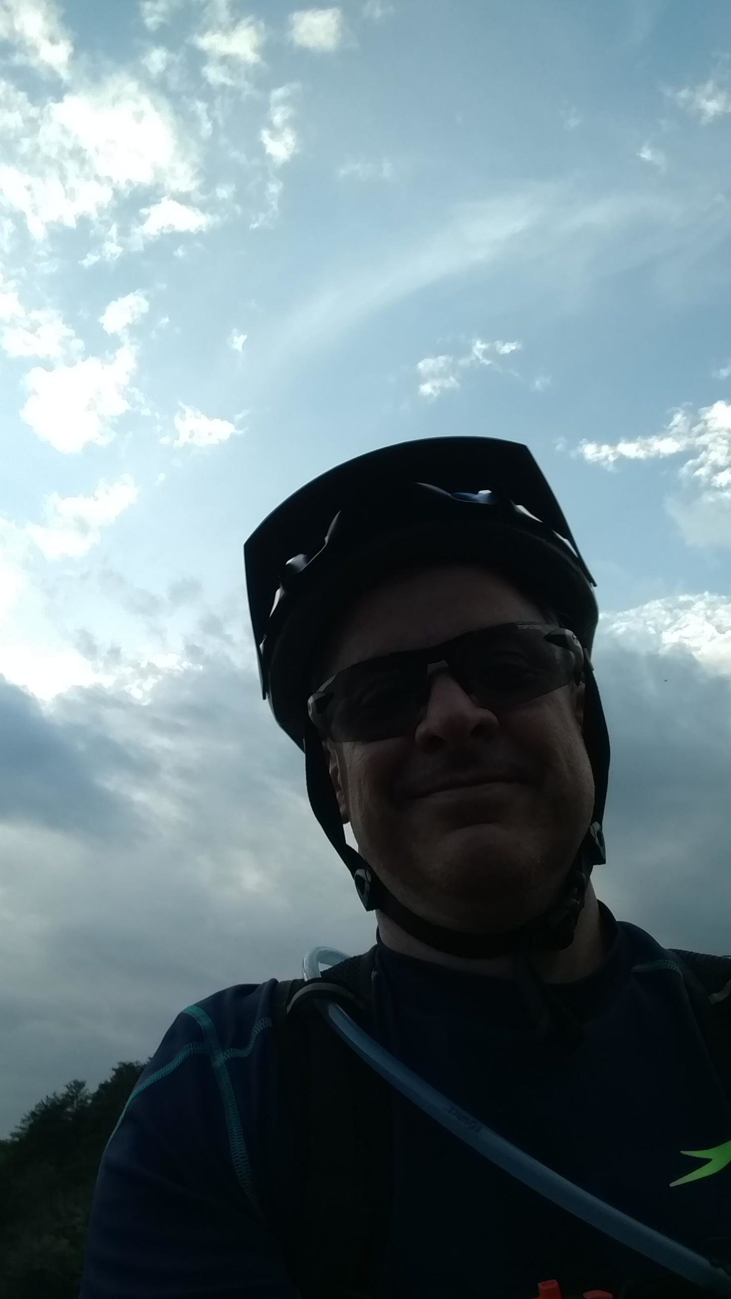 A person wearing a helmet and sunglasses smiles at the camera, with a blue sky and clouds visible in the background. The individual is dressed in a dark shirt with a light blue design and appears to be outdoors, possibly on a bike ride. Paris Mountain State Park mountain bike trail.