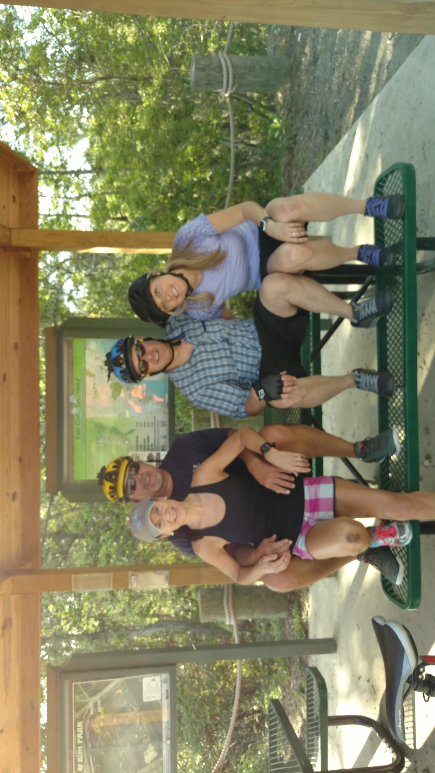 Four friends posing together on a green picnic table under a wooden shelter in a park, wearing cycling helmets and athletic clothing. The background features trees and a park information sign. Horry County Bike Run Park mountain bike trail.