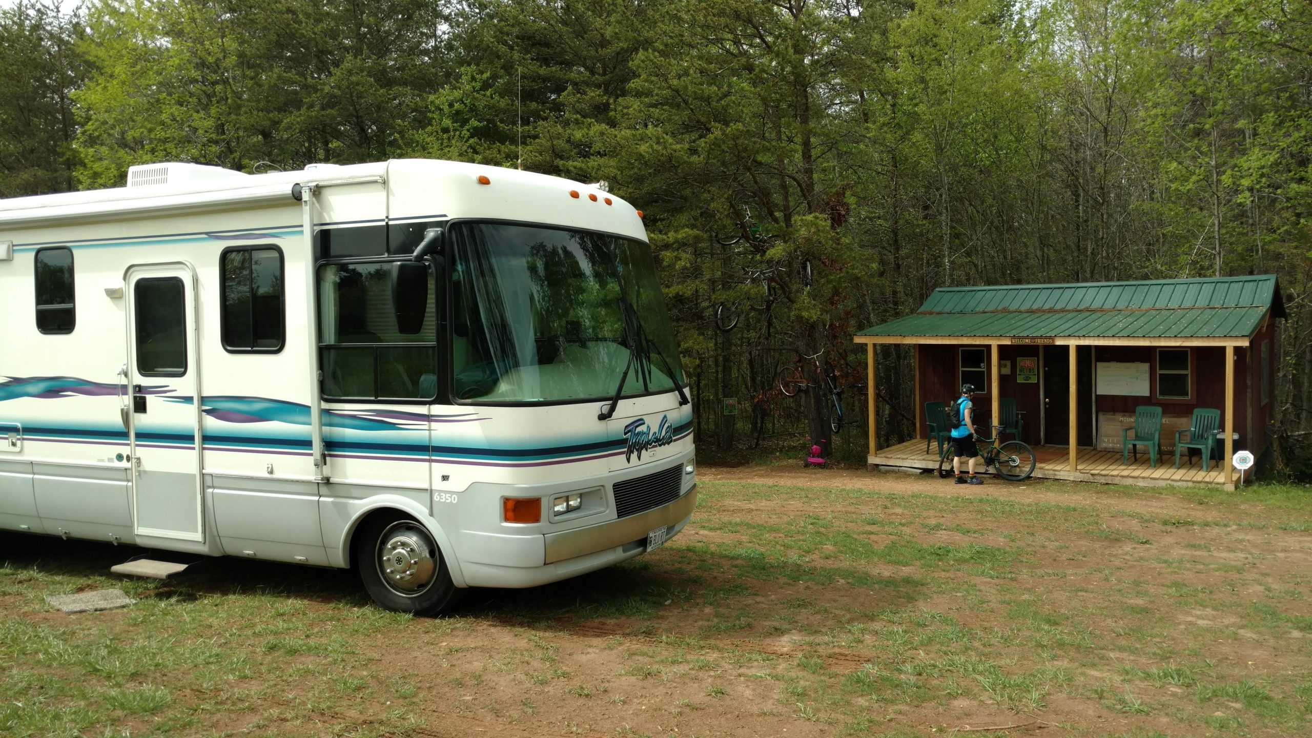 An RV parked on grass near a wooden cabin in a wooded area. A person in athletic clothing is seen standing next to a bicycle on the porch of the cabin. Trees surround the area, creating a natural backdrop. Mountain Laurel Trails mountain bike trail.