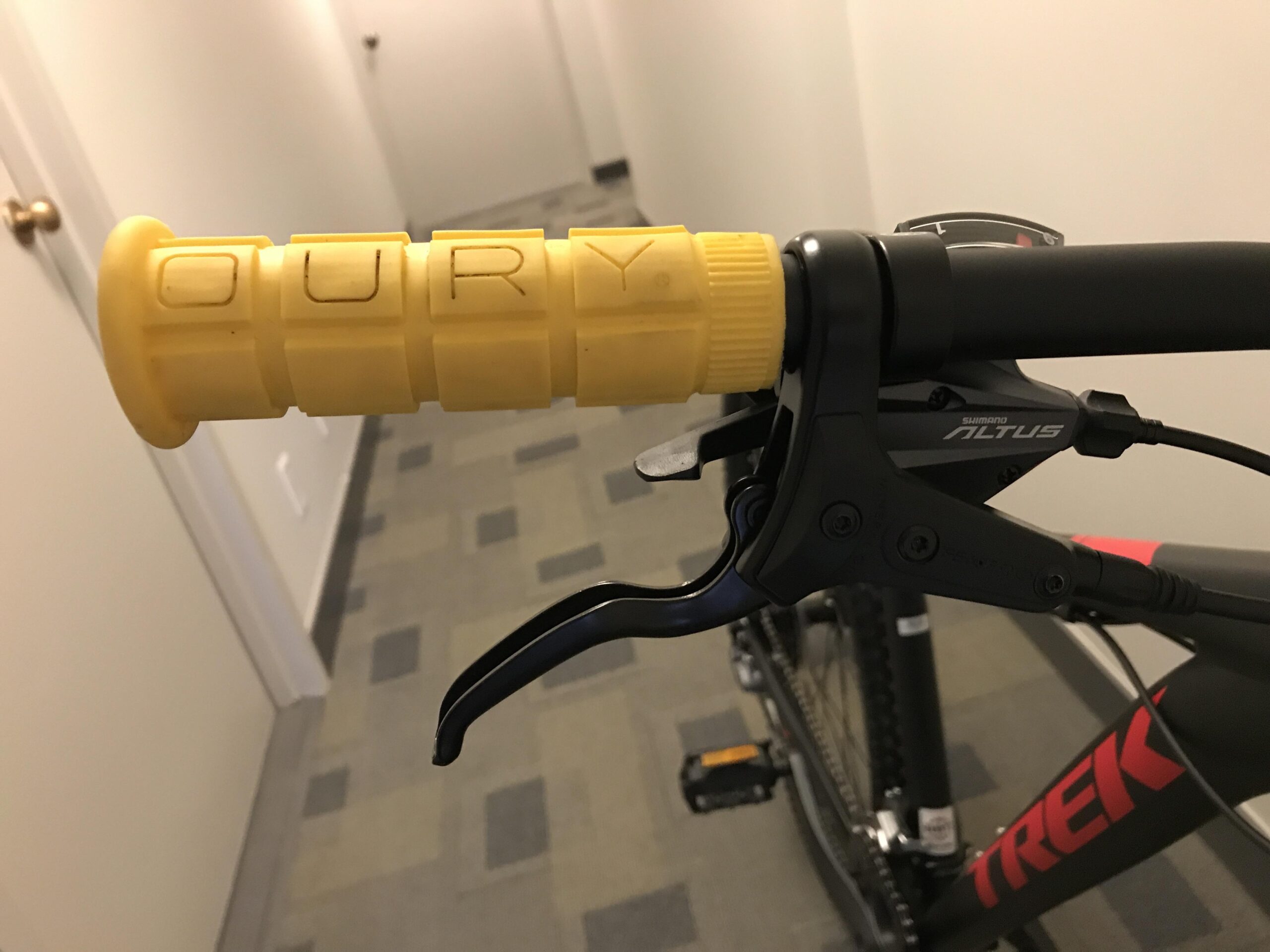 Trek 2017 Trek Marlin 7: Close-up view of a bicycle's handlebar grip, featuring a yellow Oury grip and a Shimano Altus brake lever. The background shows a hallway with neutral-colored walls and a tiled floor.