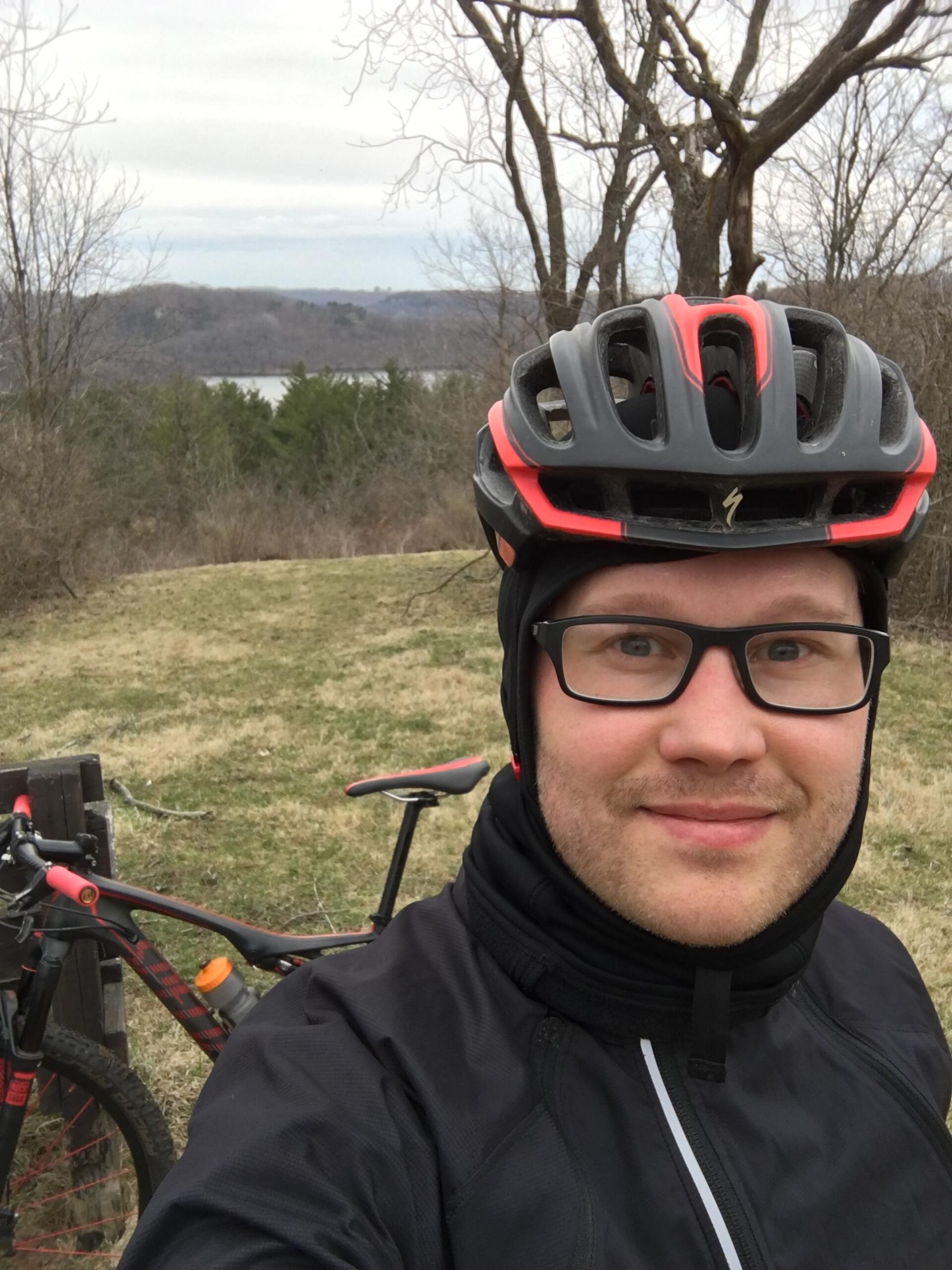 Specialized Epic Expert Carbon World Cup: A person wearing a black and red cycling helmet and winter gear stands smiling in front of a mountain bike, with a scenic view of a lake and trees in the background. The sky is overcast, hinting at a cool day.