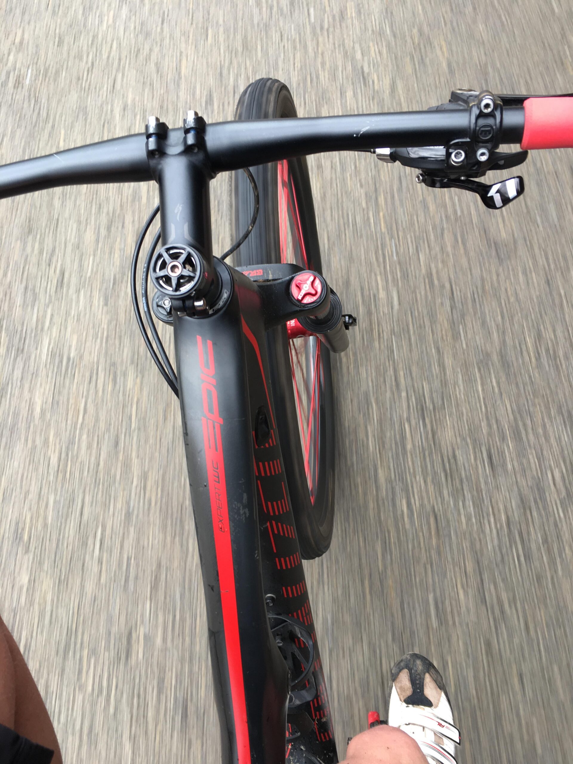 Specialized Epic Expert Carbon World Cup: A top-down view of a cyclist's handlebars and front tire while riding on a paved surface. The bike features a black and red design, with visible gear controls on the handlebars. The cyclist is wearing cycling shoes with white and black accents. The background shows a blurred road, indicating motion.