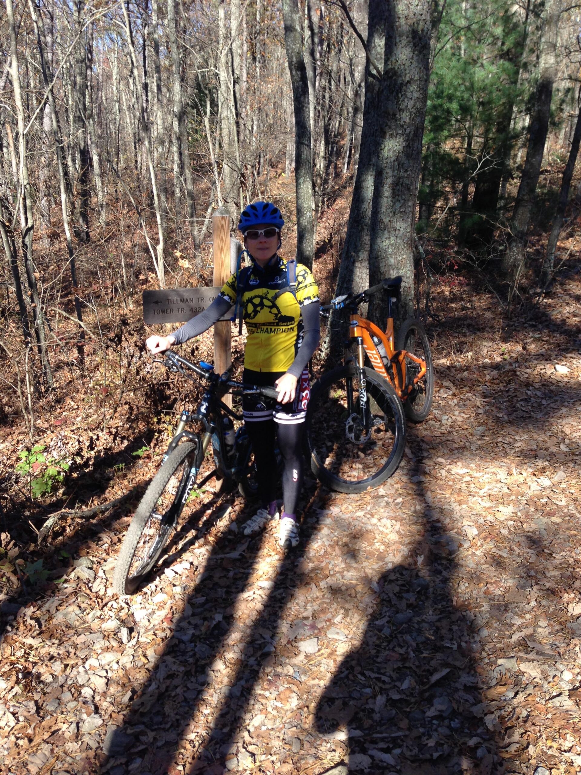 Giant Lust 2: A person in cycling gear stands beside two mountain bikes on a wooded trail covered with fallen leaves. The individual is wearing a yellow and black jersey, sunglasses, and a helmet, positioned near a wooden sign marking the trail. The background features trees and underbrush typical of a forest environment.