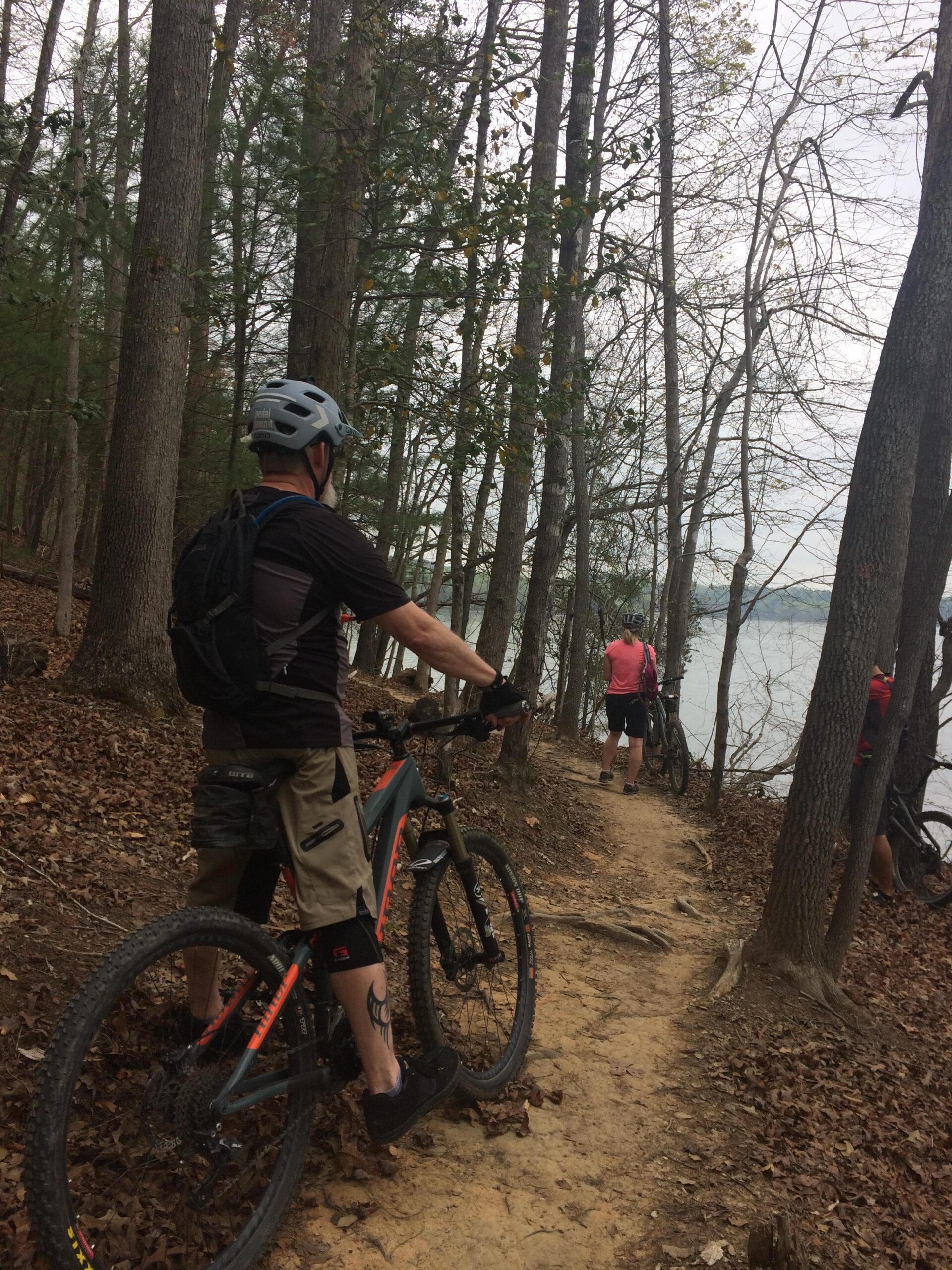 Niner RIP 9: A group of mountain bikers stops on a wooded trail along a lake. One cyclist in the foreground, wearing a helmet and casual biking attire, stands next to his bike, while two others are further down the path. Surrounding them are tall trees and fallen leaves, with a cloudy sky overhead.