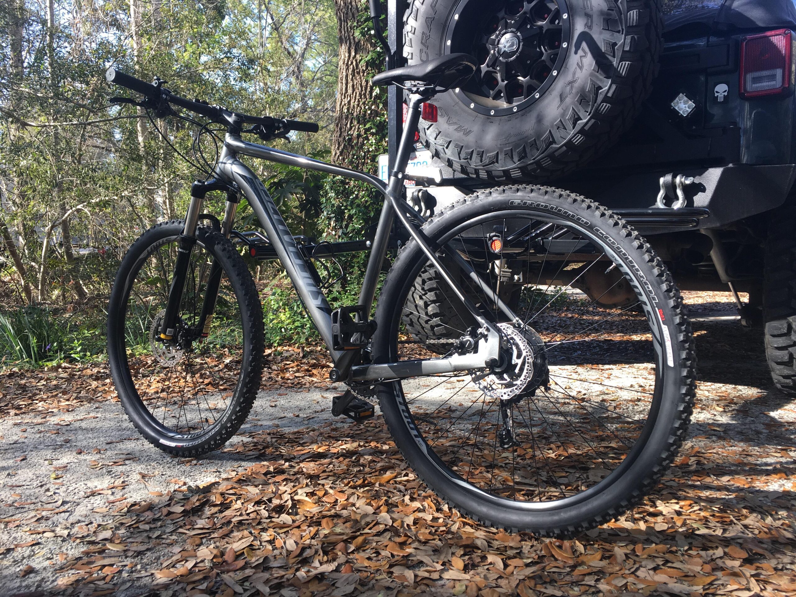 Specialized Rockhopper Comp 29: A black mountain bike positioned next to a parked vehicle, surrounded by fallen leaves on a gravel surface. The background features trees and greenery, indicating a natural outdoor setting. The bike has a sleek design with thick tires and a front suspension fork.