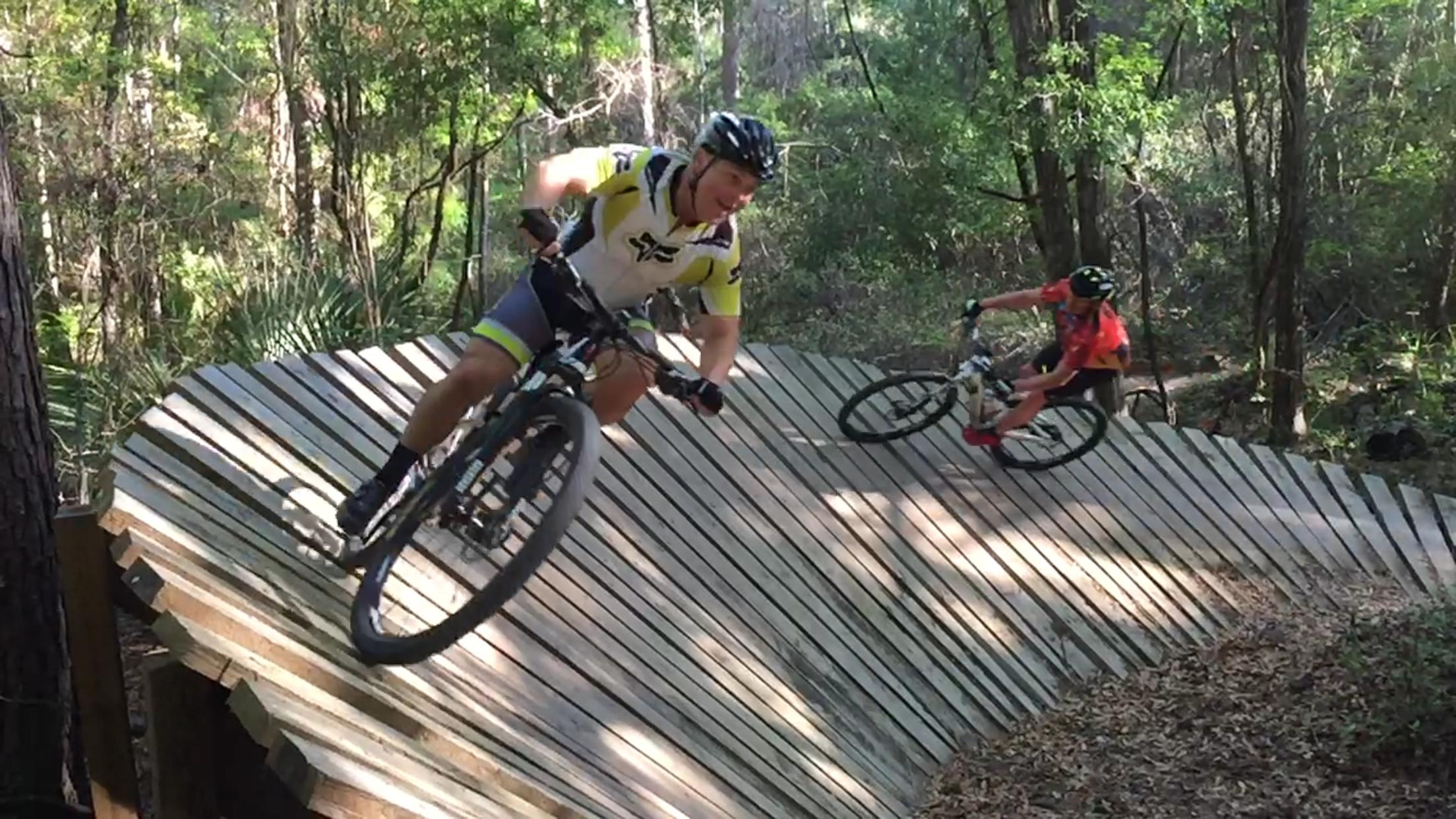 Two mountain bikers navigate a wooden ramp in a forested area. One rider, wearing a yellow and black jersey, leans into a turn, while the other, in an orange shirt, follows closely behind. Lush greenery surrounds the scene, highlighting the outdoor adventure. Santos mountain bike trail.