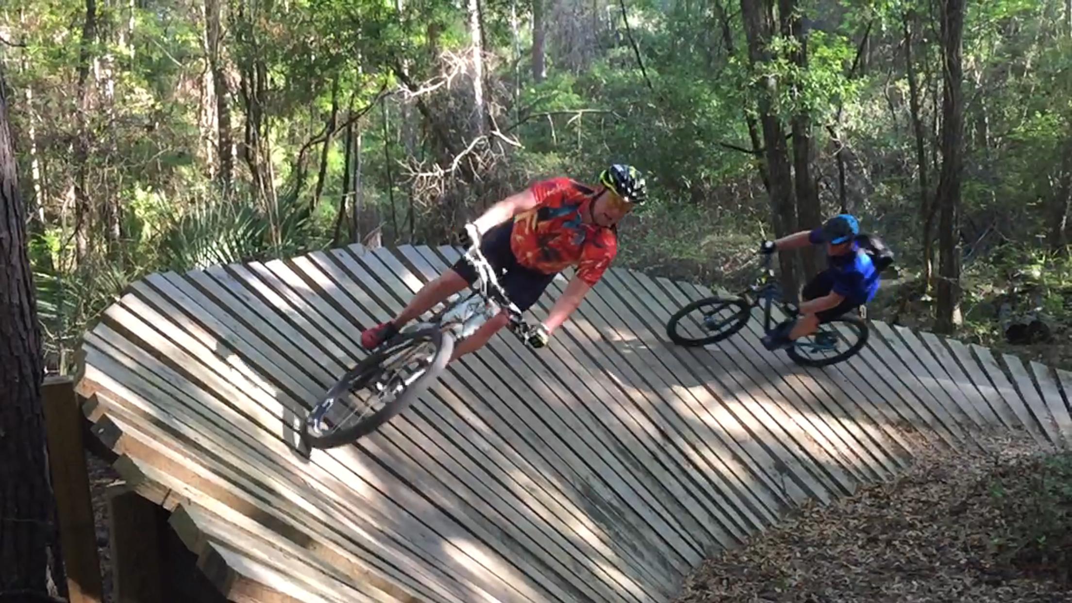 Two mountain bikers navigate a wooden pump track in a lush forest. One rider leans into a turn while the other closely follows, showcasing their skills on the curved boards of the track. Sunlight filters through the trees, creating a vibrant atmosphere in the natural setting. Santos mountain bike trail.
