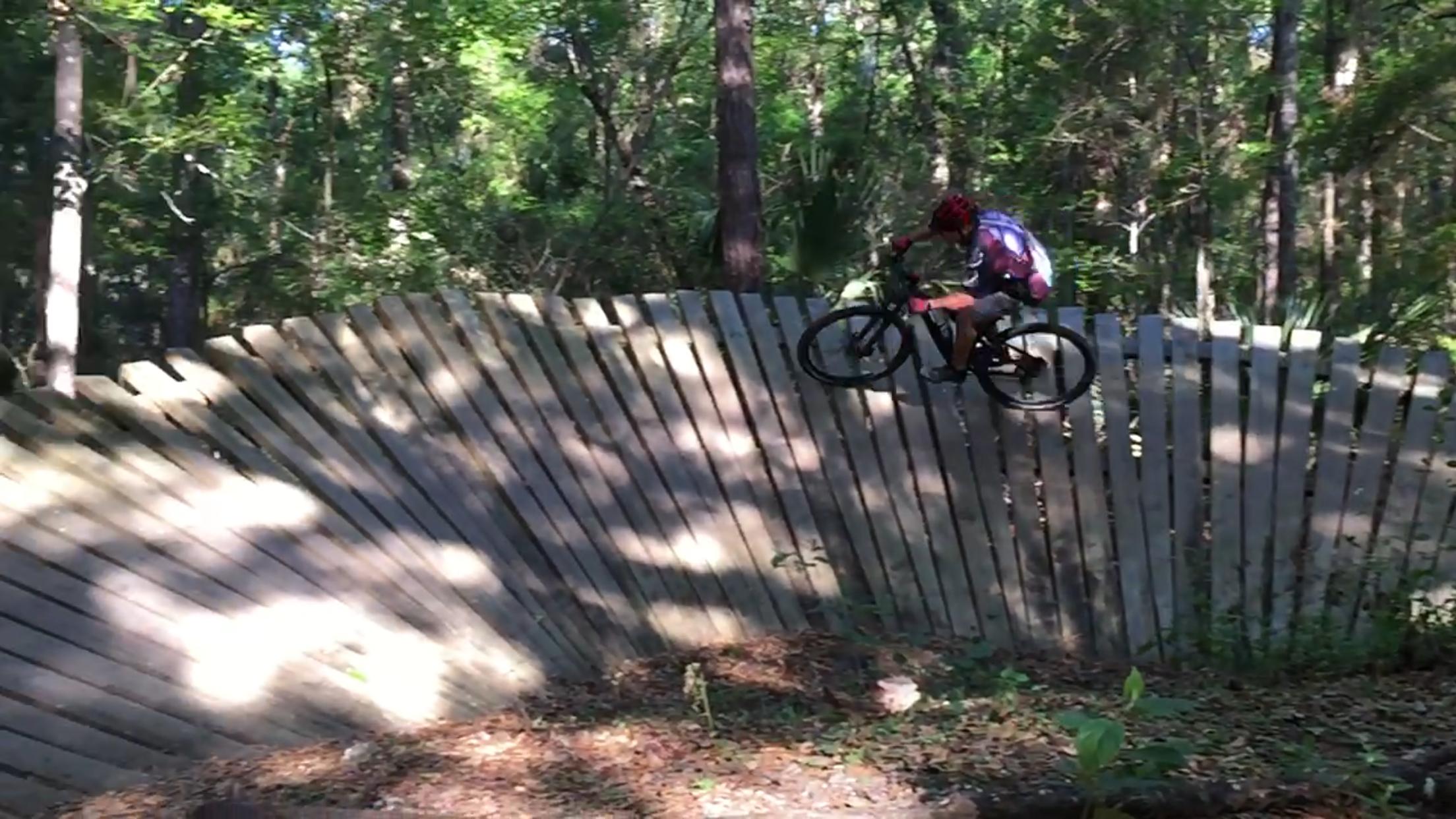 A cyclist performing a jump on a wooden ramp in a forested area, surrounded by green trees and foliage. The cyclist is captured mid-air with a focused expression, showcasing a dynamic mountain biking scene. Santos mountain bike trail.