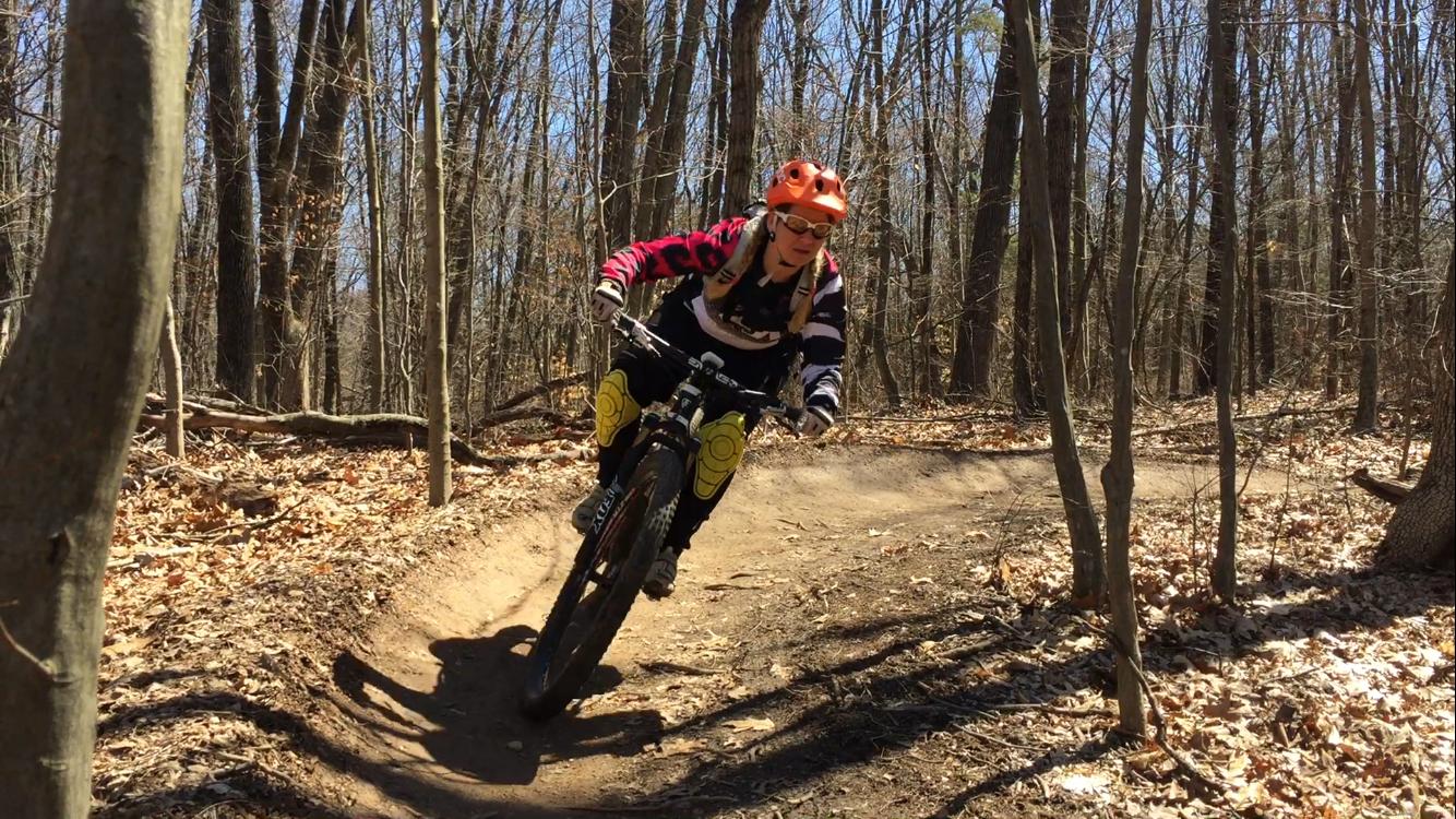 A mountain biker navigates a twisty trail in a wooded area, surrounded by tall trees and fallen leaves. The rider wears an orange helmet, sunglasses, and a colorful long-sleeve jersey, demonstrating skill as they lean into a turn on their bike. The scene captures the essence of outdoor adventure and sports. Nockamixon State Park mountain bike trail.