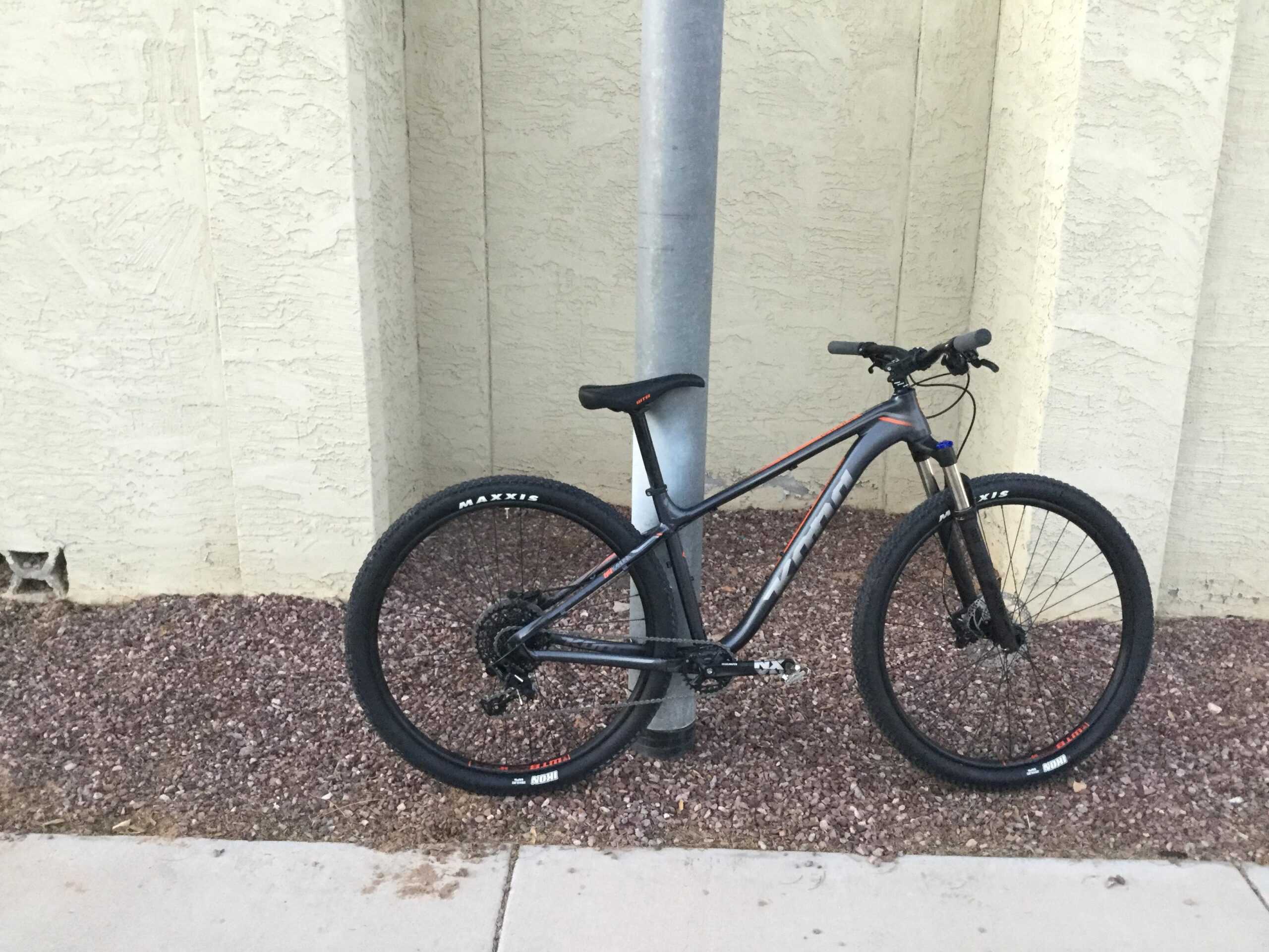 Kona Kahuna: A mountain bike resting against a utility pole, positioned on a gravel surface next to a sidewalk, with a textured yellow wall in the background. The bike features large black tires and a sleek frame design.