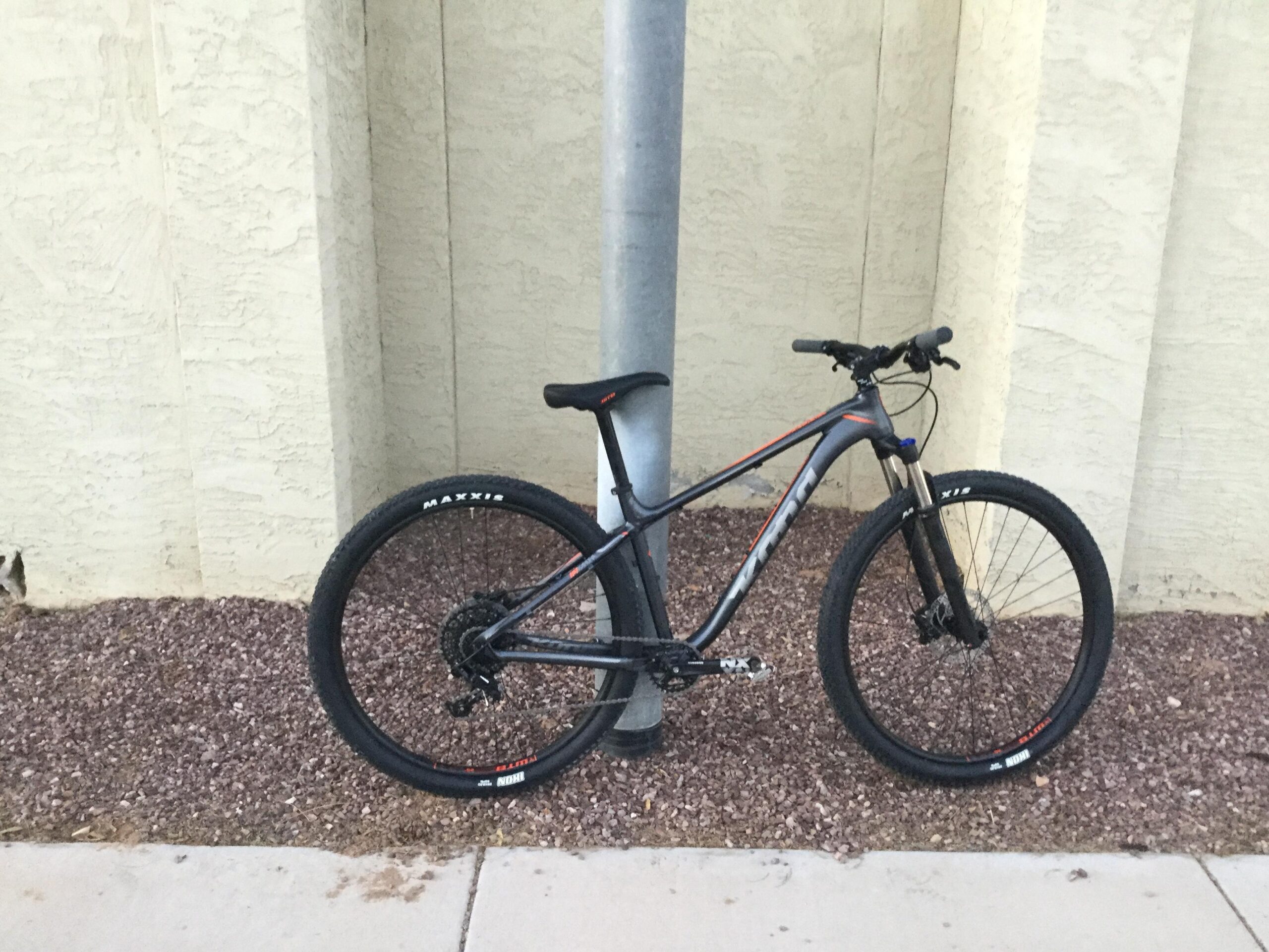 Kona Kahuna: A mountain bike leaning against a street pole, positioned in front of a textured, light-colored wall. The bike has black tires with red and white accents and features a sleek frame design. Small gravel is scattered on the ground near the bike.