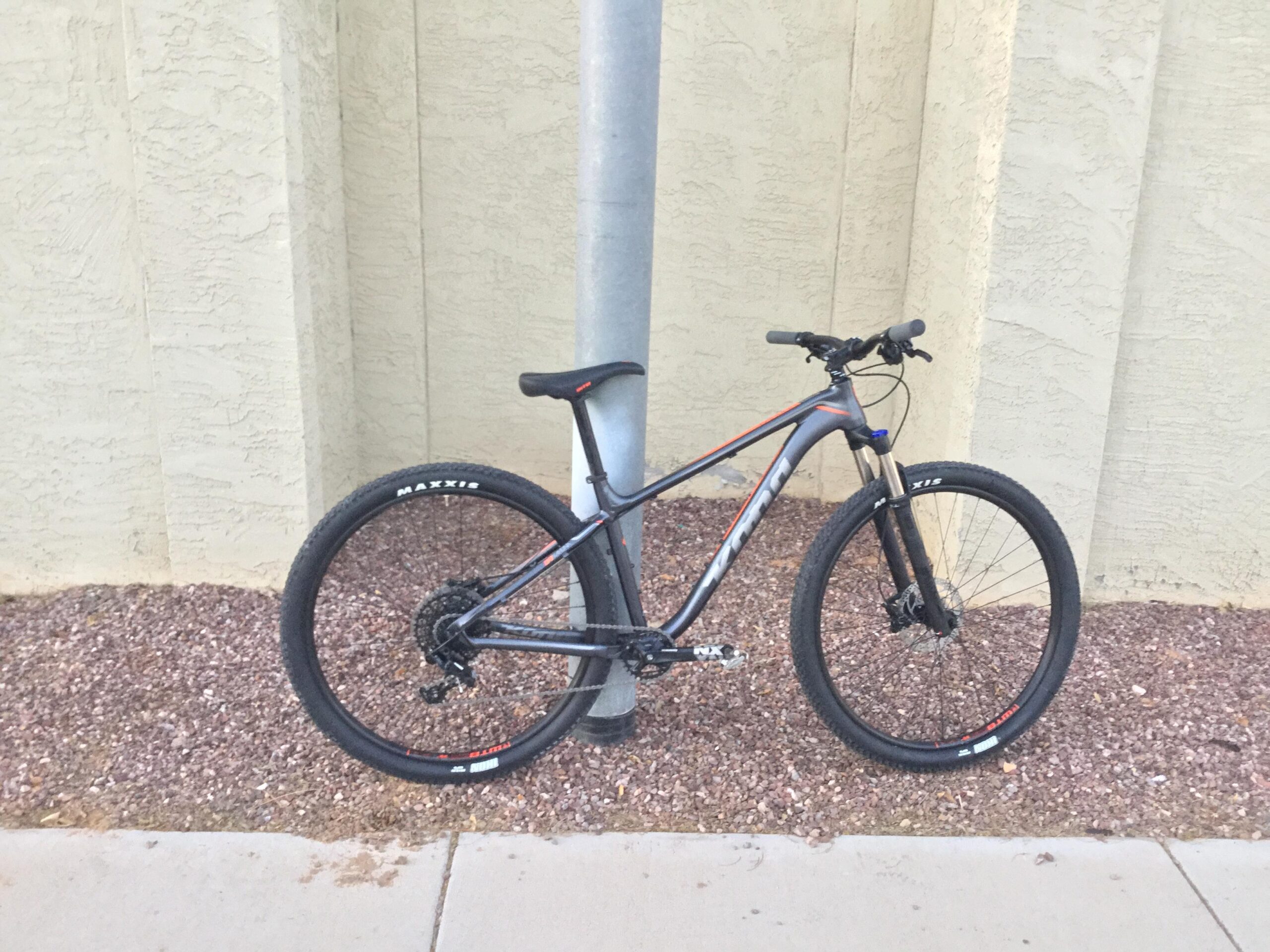 Kona Kahuna: A mountain bike leaning against a pole, with a background of textured wall and gravel.