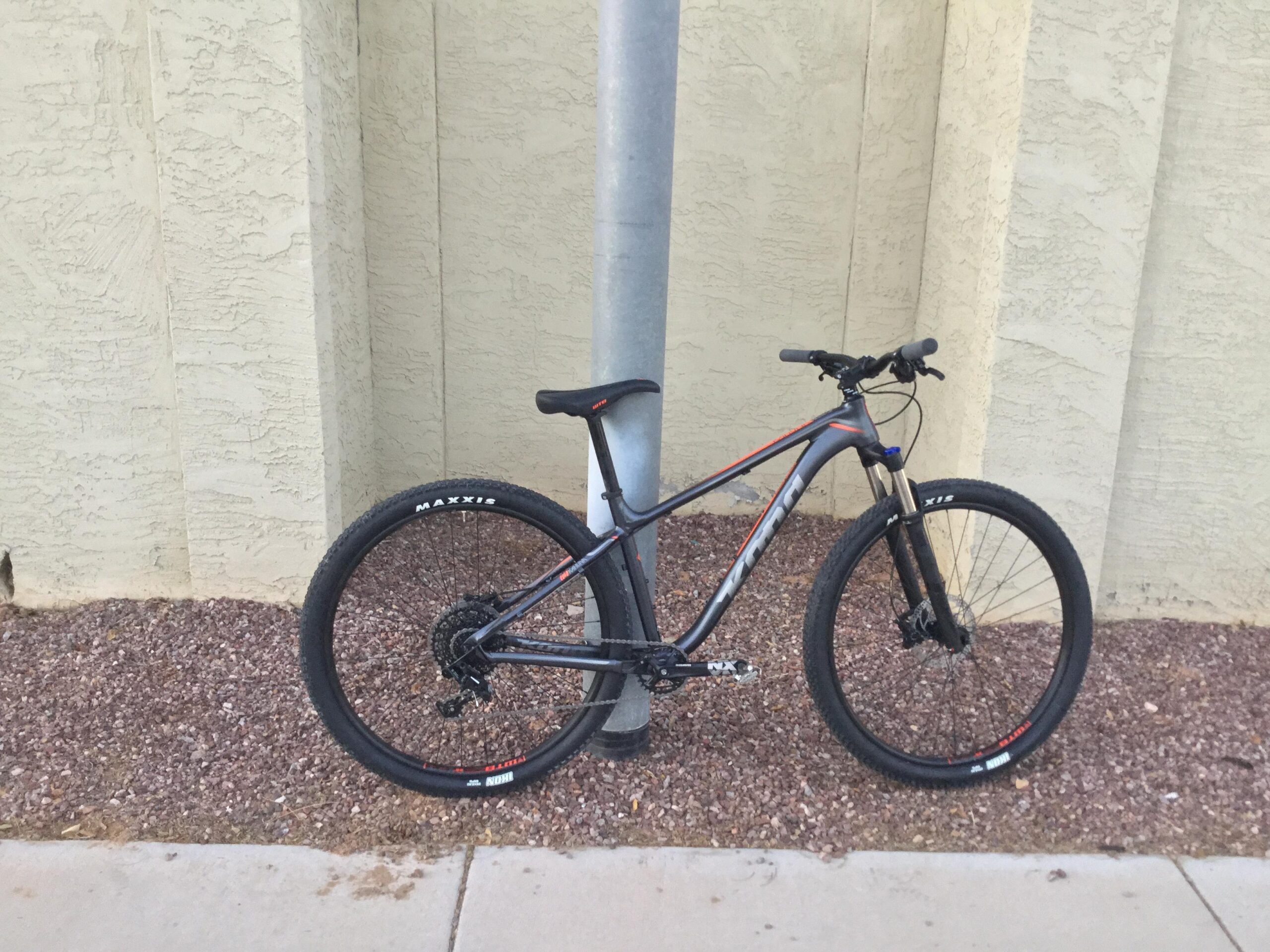 Kona Kahuna: A mountain bike leaning against a utility pole, positioned next to a light-colored textured wall. The bike is primarily black with orange accents and features thick tires with "MAXXIS" branding. The ground is covered with small gravel stones.