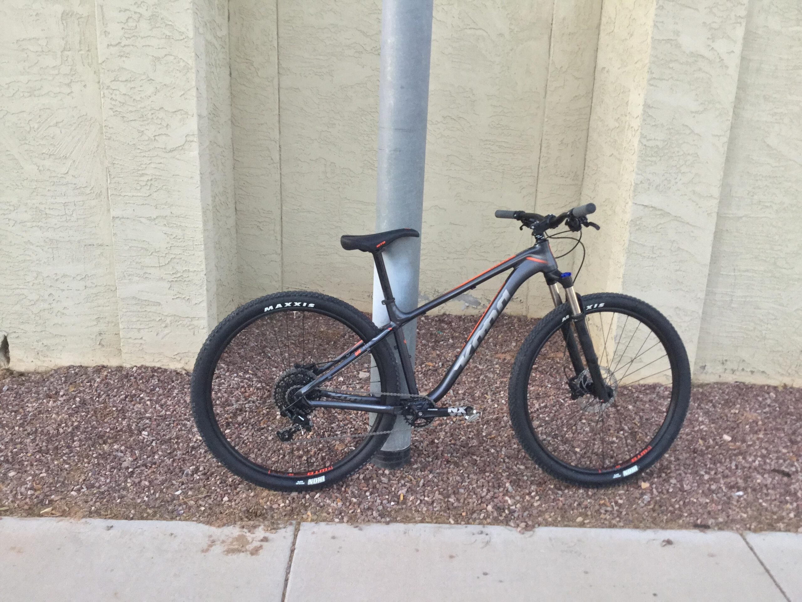 Kona Kahuna: A modern mountain bike leaning against a metal pole, positioned next to a textured beige wall. The bike features black tires with visible branding and a gray frame with orange accents. Small gravel decorates the ground surrounding the bike.